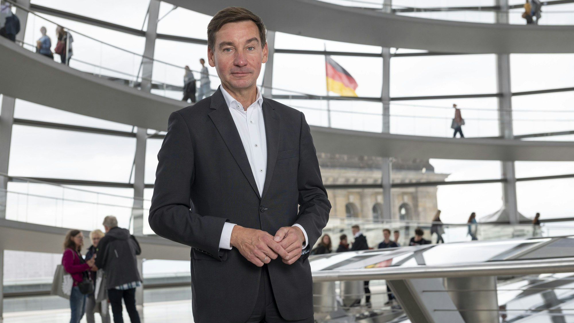 Ein Mann im Anzug steht in einem Gebäude mit großen Fenstern. Im Hintergrund weht eine Deutschlandflagge.