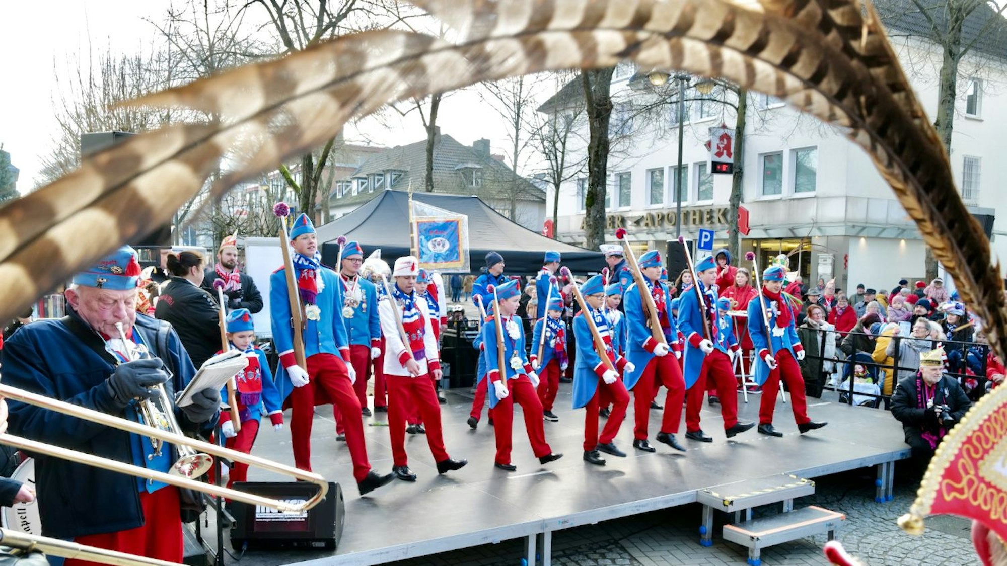 Musiker in roten Hosen und blauen Oberteilen stehen inmitten einer Stadt auf einer Plattform. Die Aussicht auf die Gruppe wird von der Feder an einem karnevalistischen Hut gerahmt.
