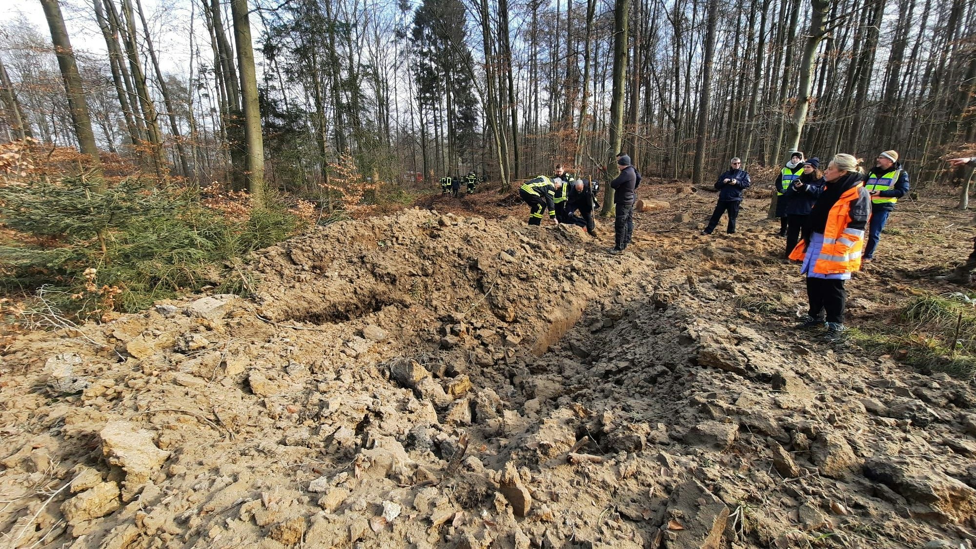 Ein Waldgelände mit einem Krater im Vordergrund; mehrere Personen stehen in der Nähe.
