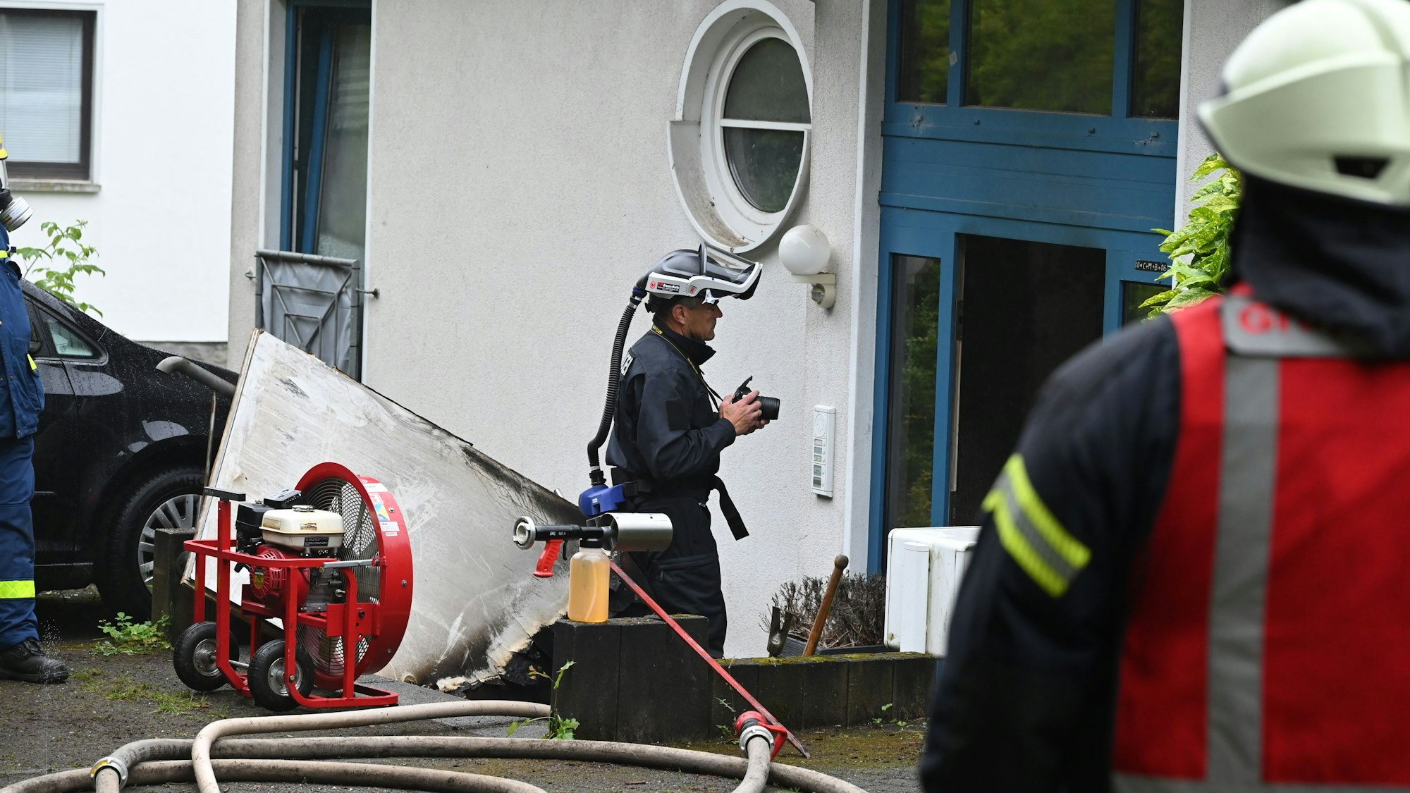 Ein Feuerwehrmann ist von hinten zu sehen. Er schaut nach einem Brand auf einen Hauseingang, vor dem ein Ermittler der Polizei arbeitet.
