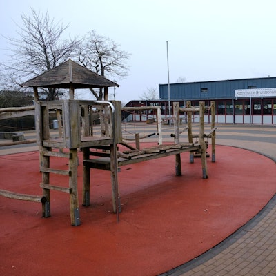 Eine Frau geht über den Pausenhof der Katholischen Grundschule in Kommern. Links im Bild ist ein Klettergerüst aus Holz zu sehen.