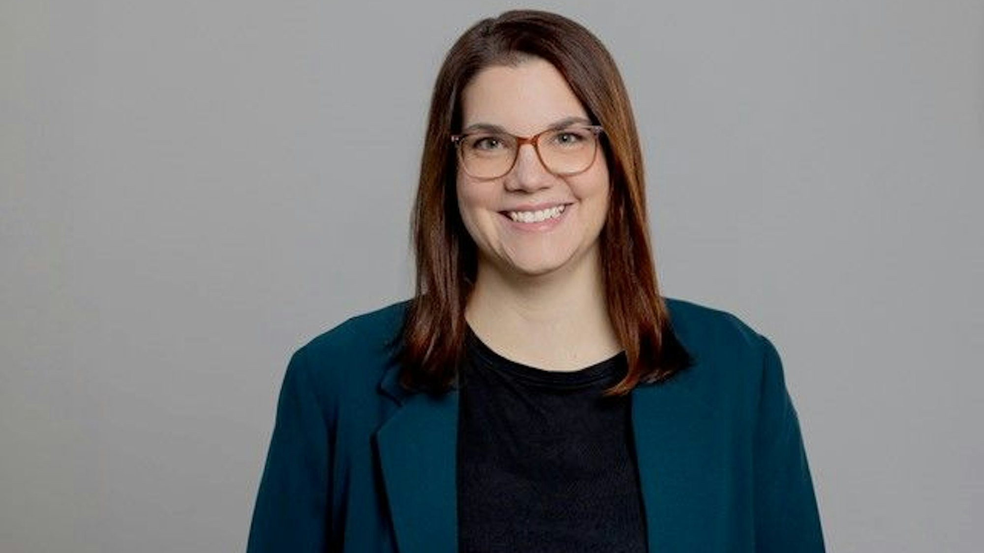 Eine Frau mit Brille und Sakko grinst vor einem grauen Hintergrund in die Kamera.