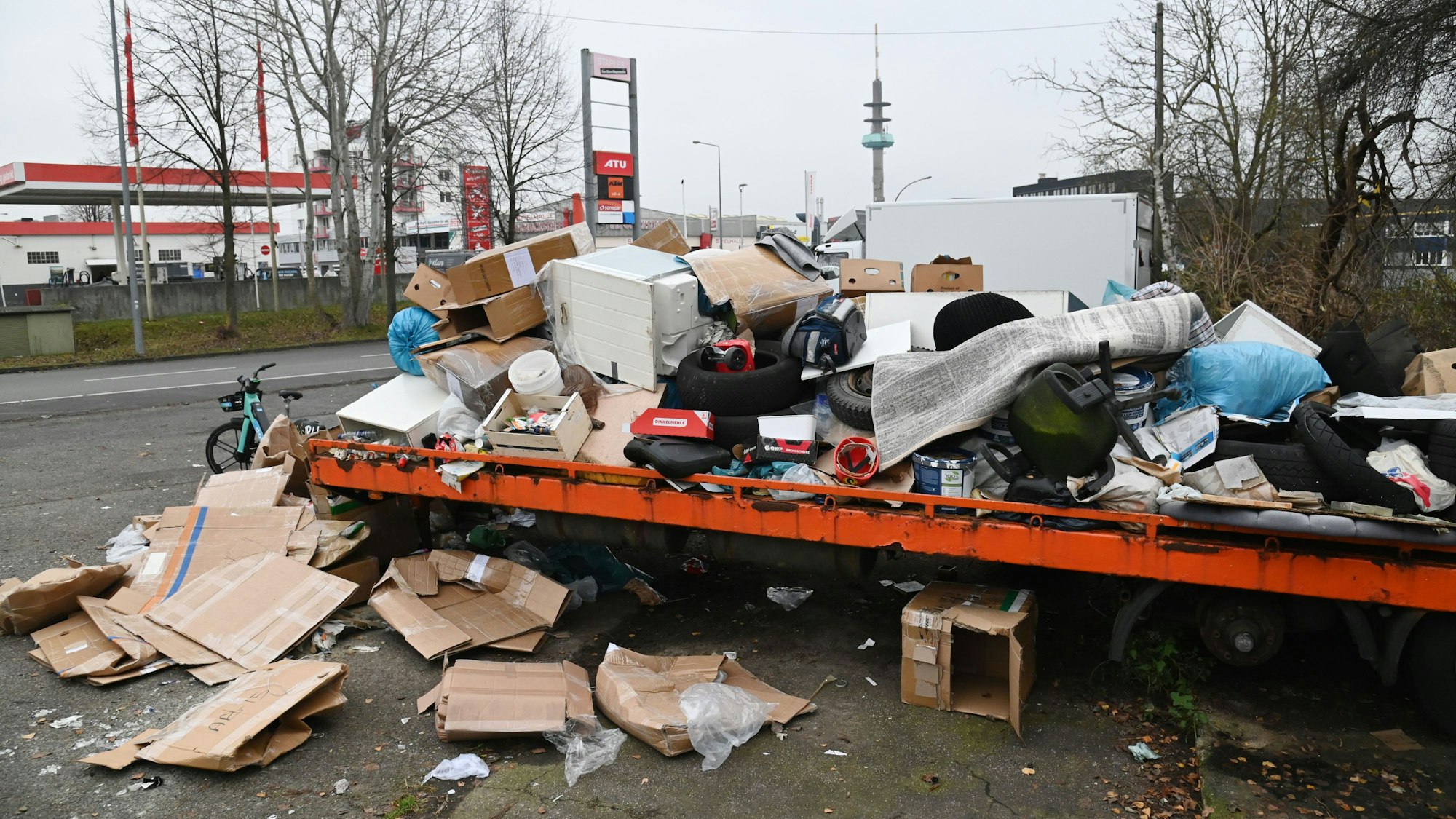 Am Rande einer Straße hat sich auf einem Lkw-Anhänger eine große Menge wild abgeladenen Mülls angesammelt: ausrangierte Möbel, Teppiche, Plastiksäcke und Kartons aus Pappe sind übereinander getürmt und verunreinigen zum Teil den Gehweg.