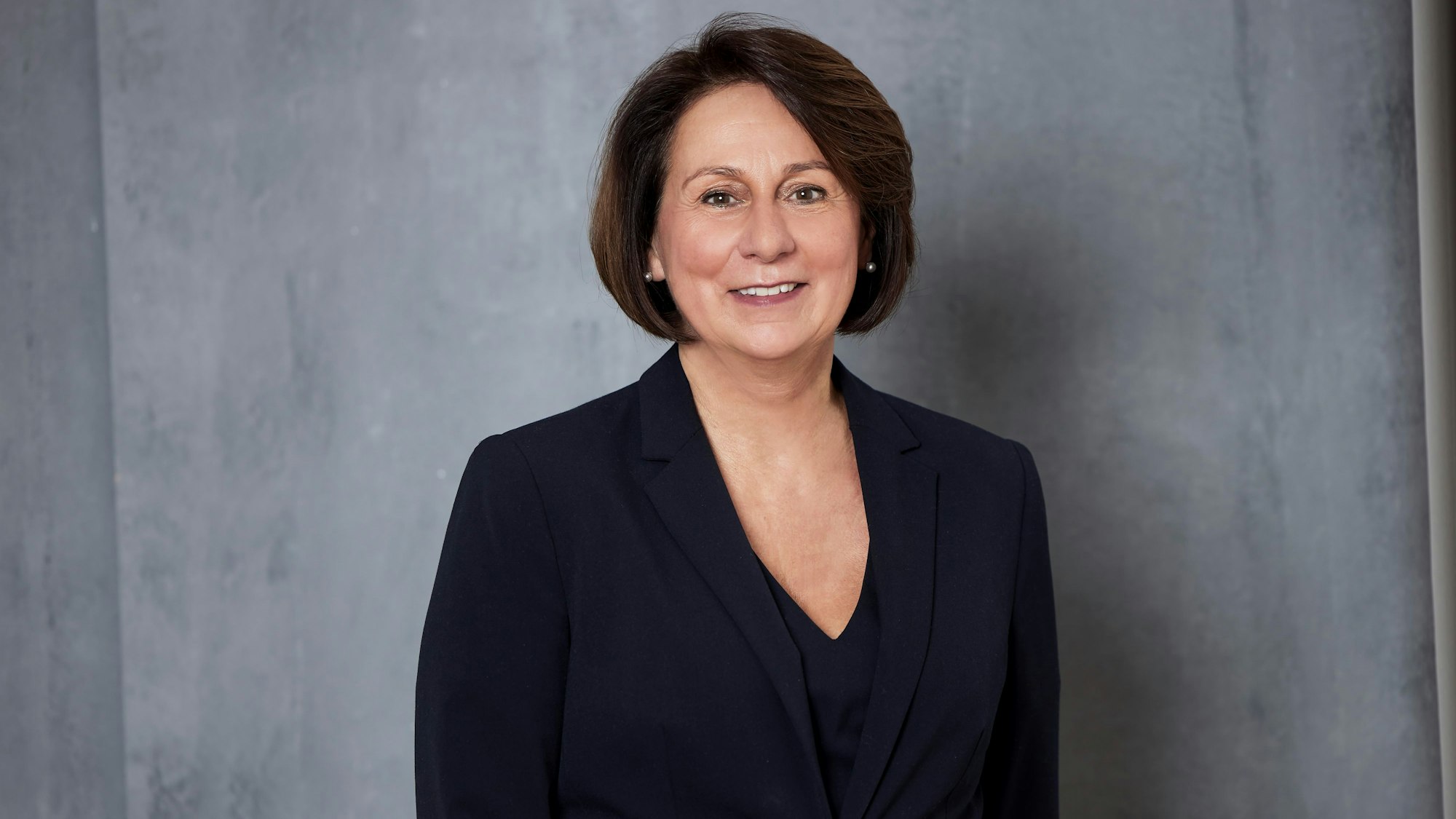 Eine Frau in einem Anzug steht vor einer grauen Wand.