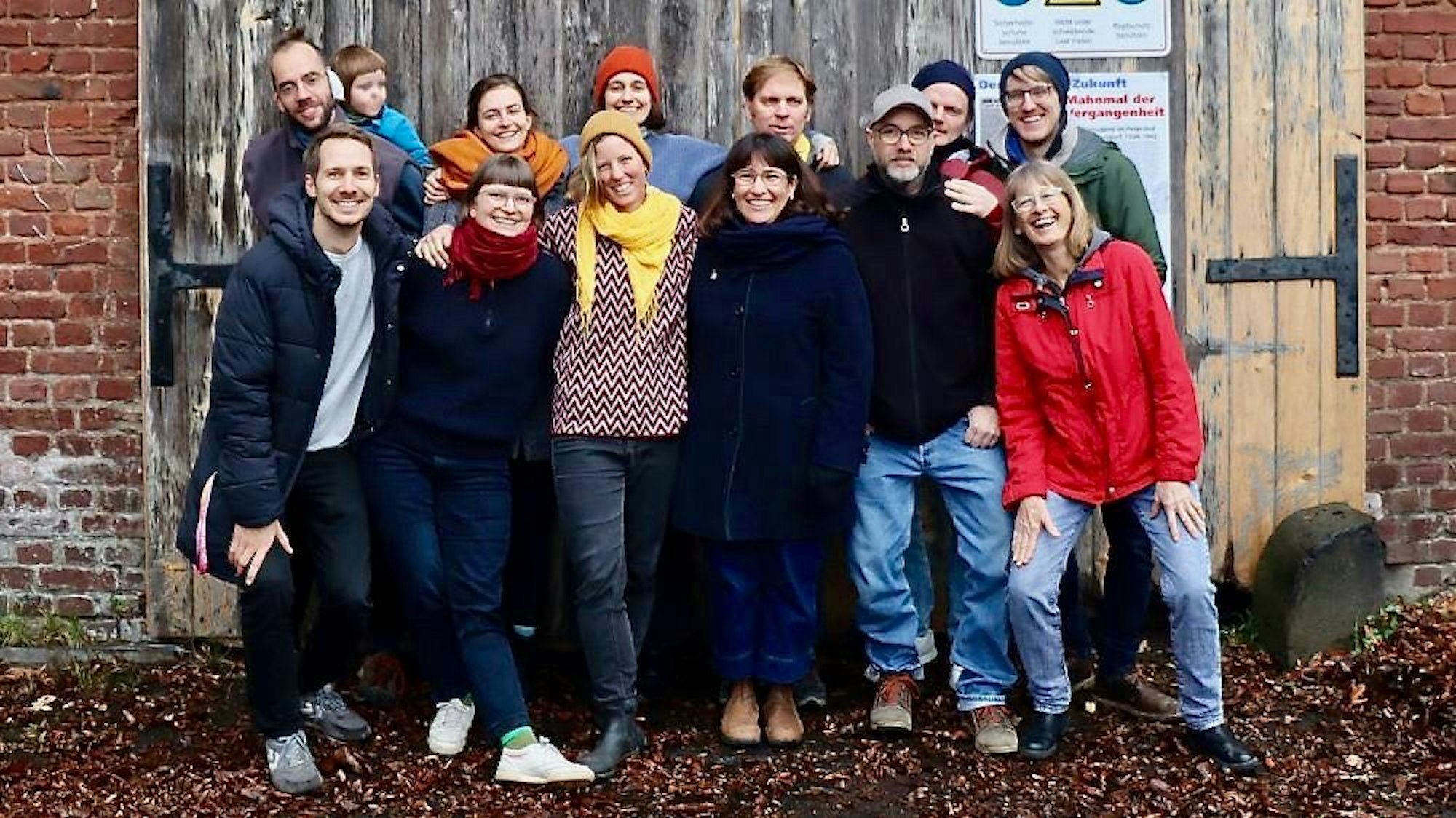 Vor einem hölzernen Tor in einer Ziegelmauer steht eine Gruppe Menschen.