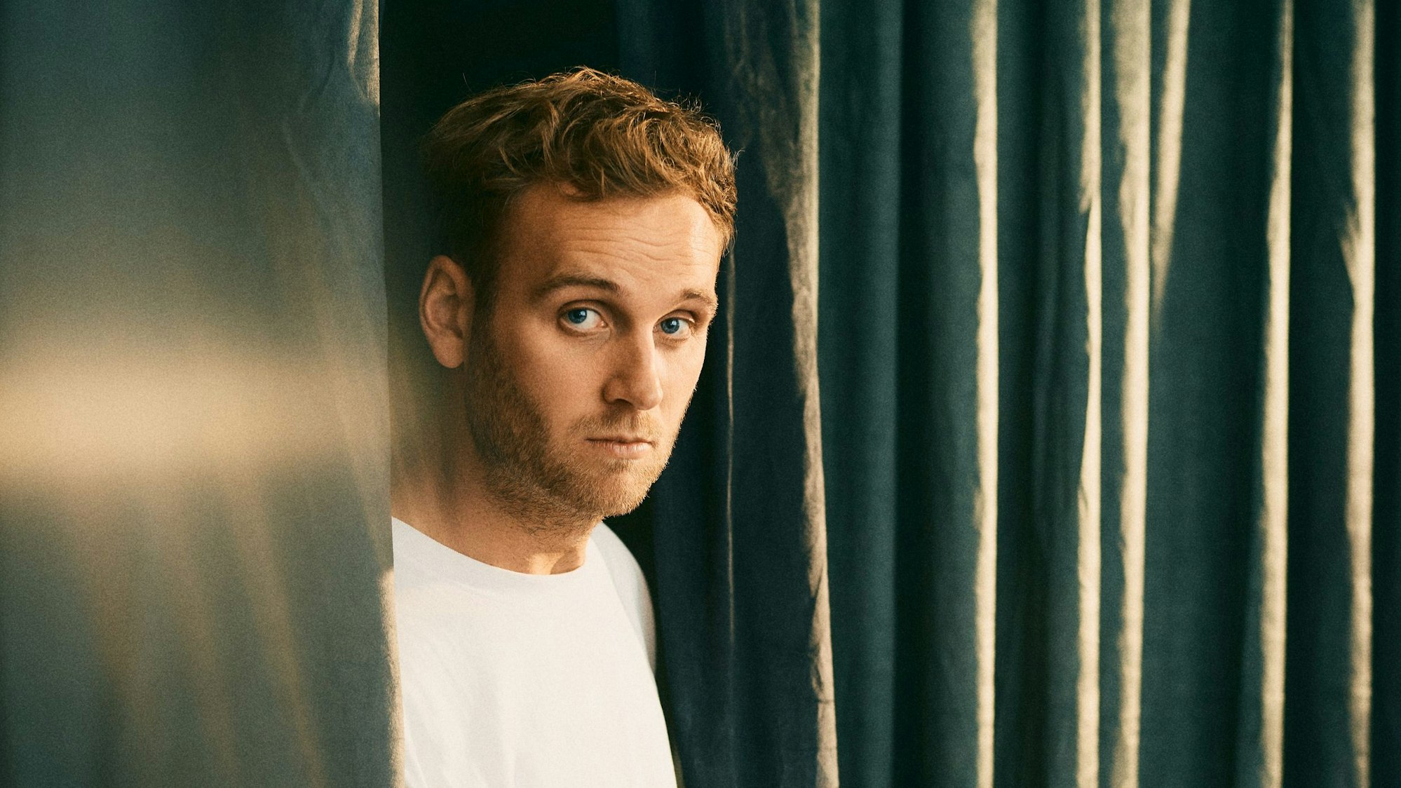 Portrait von Leon Windscheid, während er zwischen einem grünen Vorhang steht.