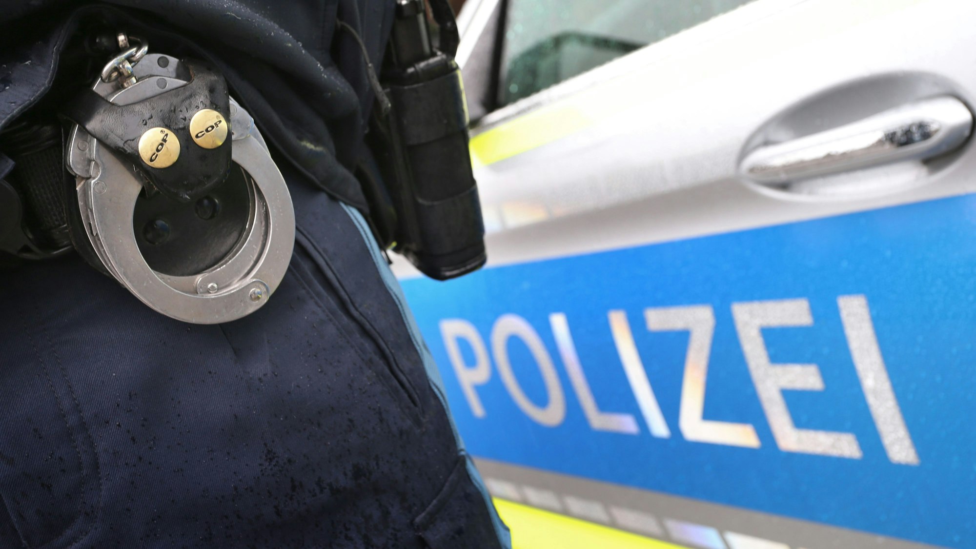 Ein Polizist mit Handschellen und einer Pistole am Gürtel steht vor einem Streifenwagen.