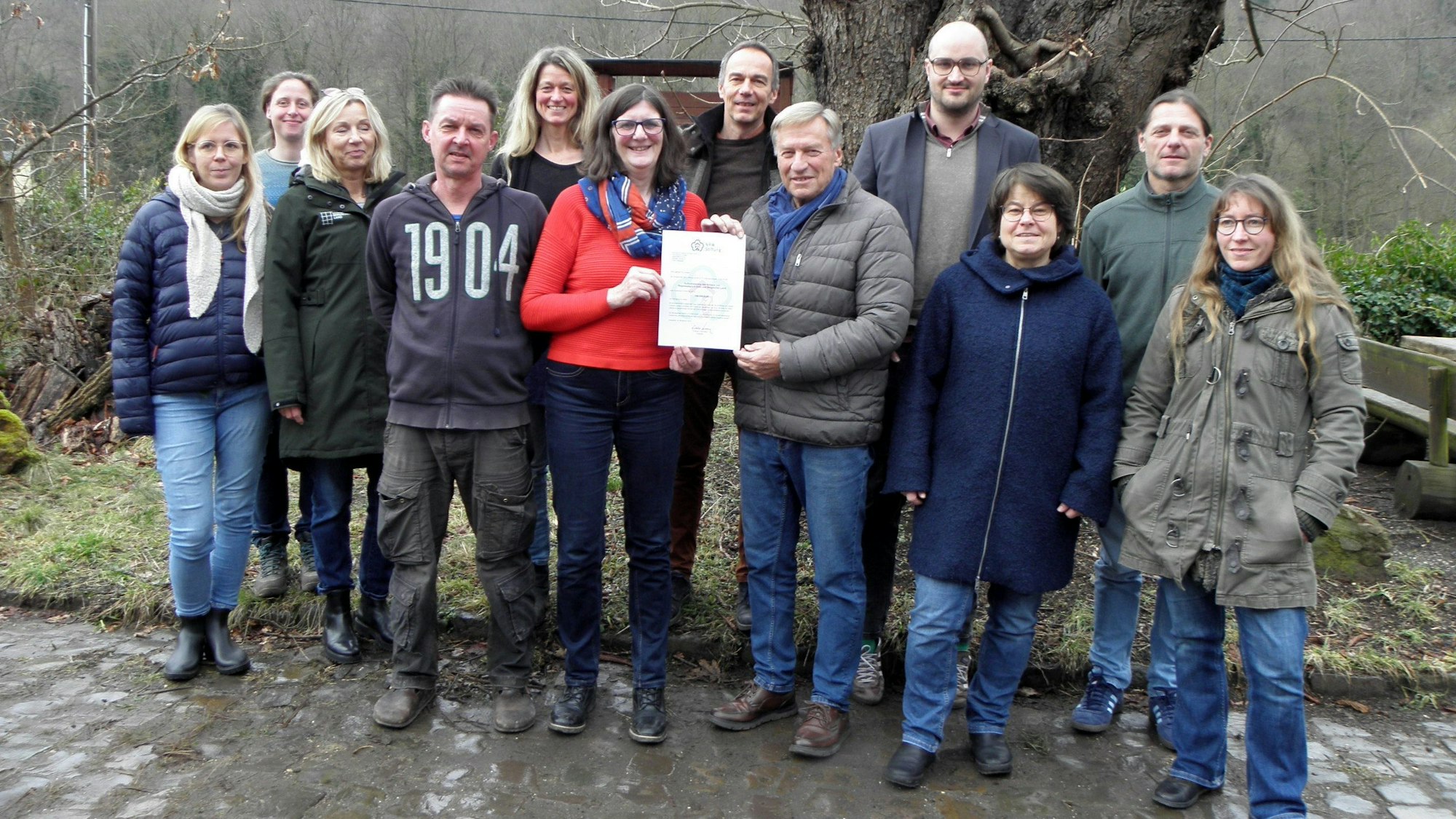 Eine Gruppe Menschen steht vor einem Baum, in der Mitte halten ein Mann und eine Frau eine Förderurkunde in die Kamera.