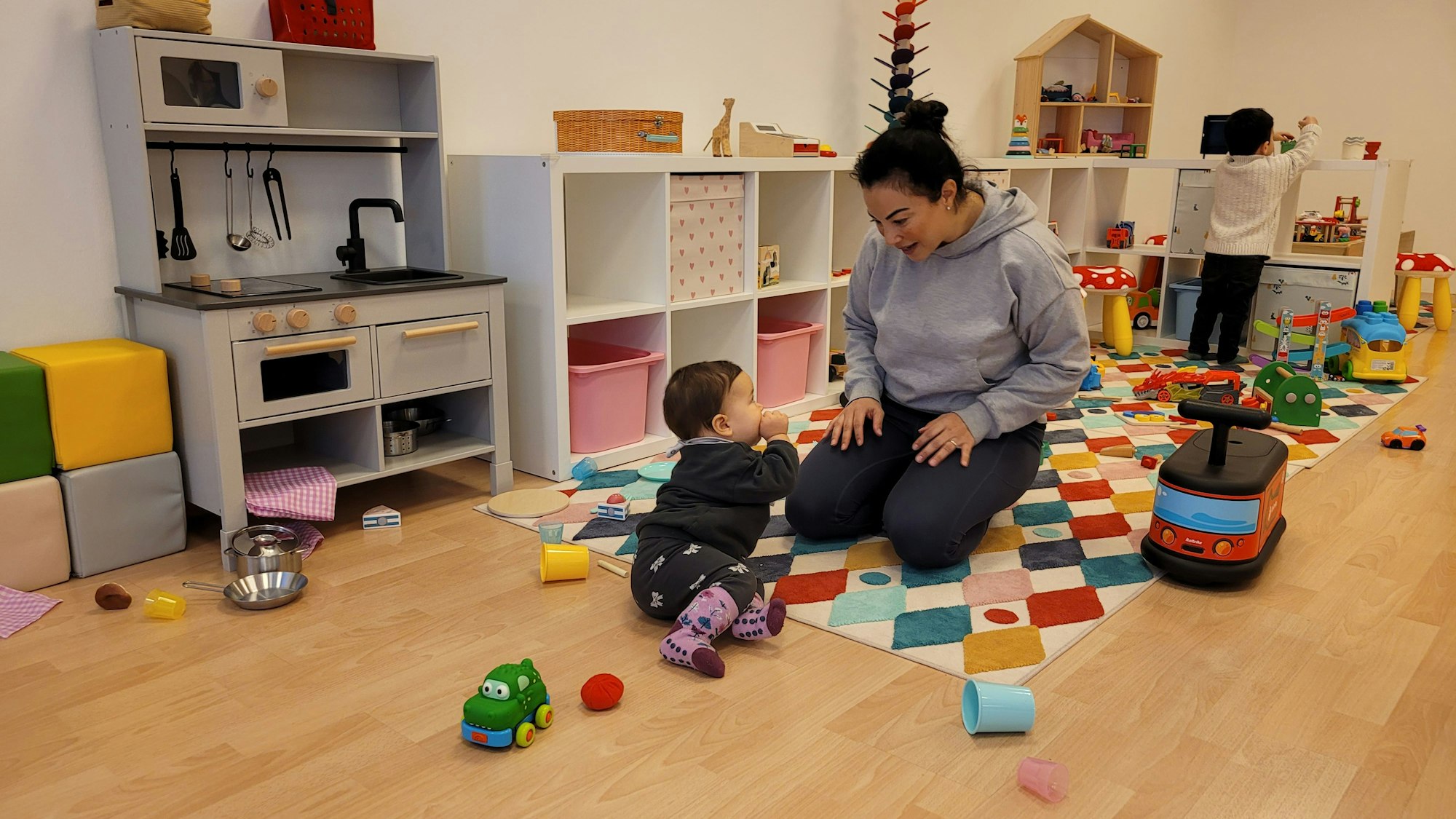 Eine Frau kniet auf einem Teppich mit bunten Rauten, vor sitzt ihre kleine Tochter.