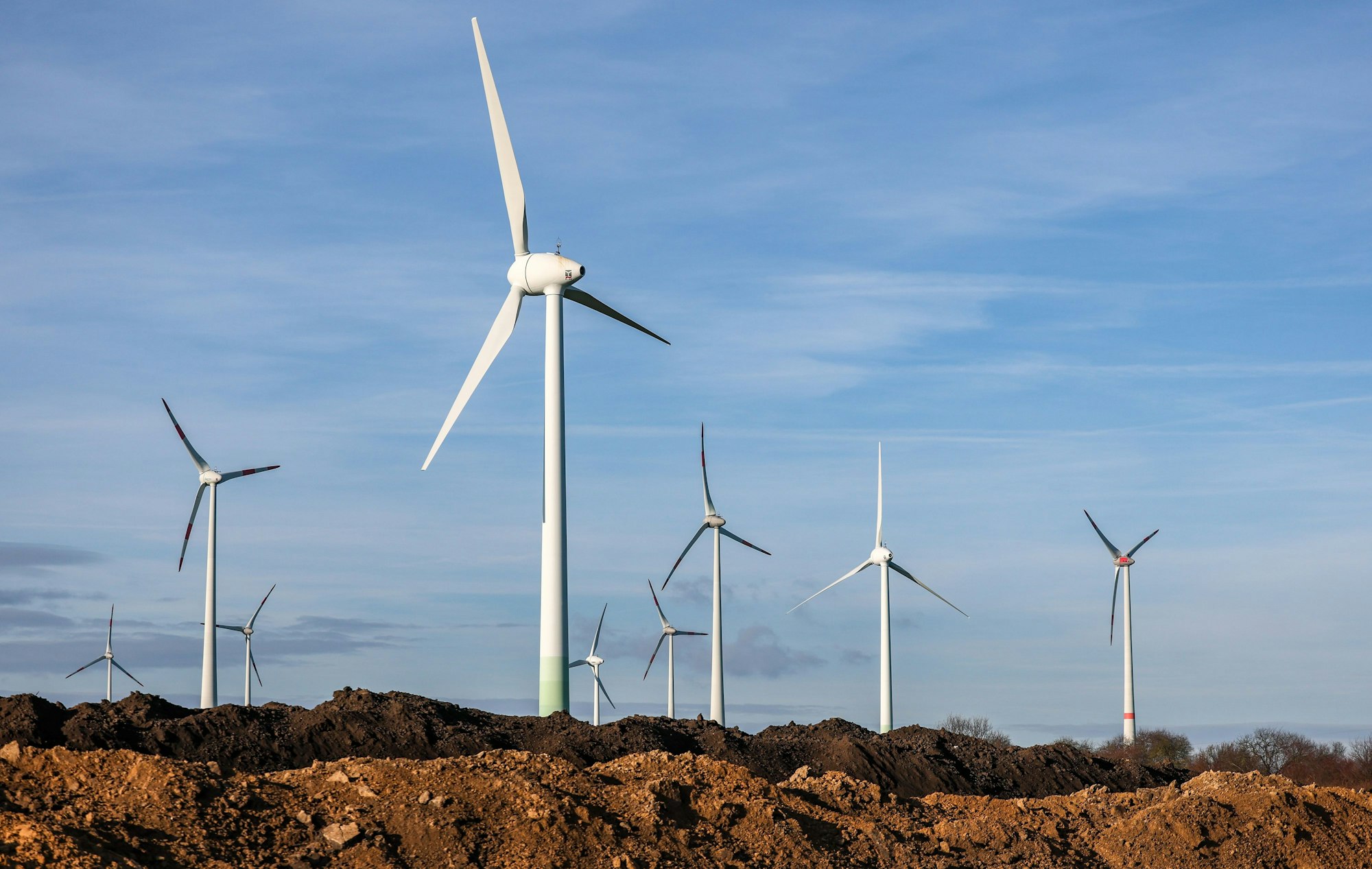 Windräder stehen hinter Erdhaufen am Rande eines Baufelds. Ihr Bau könnte durch die Zölle letztendlich günstiger werden.