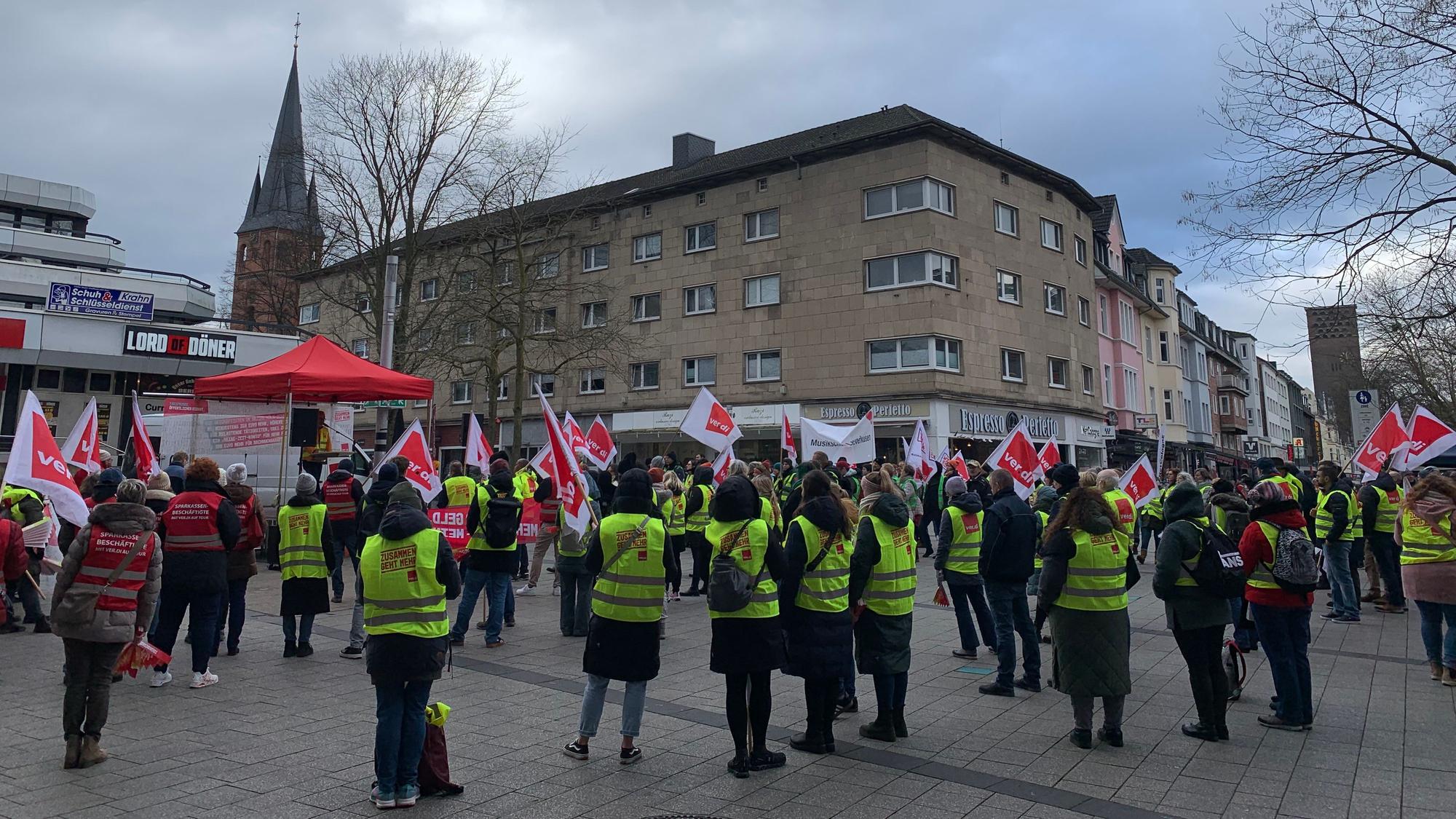 Viele Menschen stehen mit Fahnen und Warnwesten auf einem Platz.