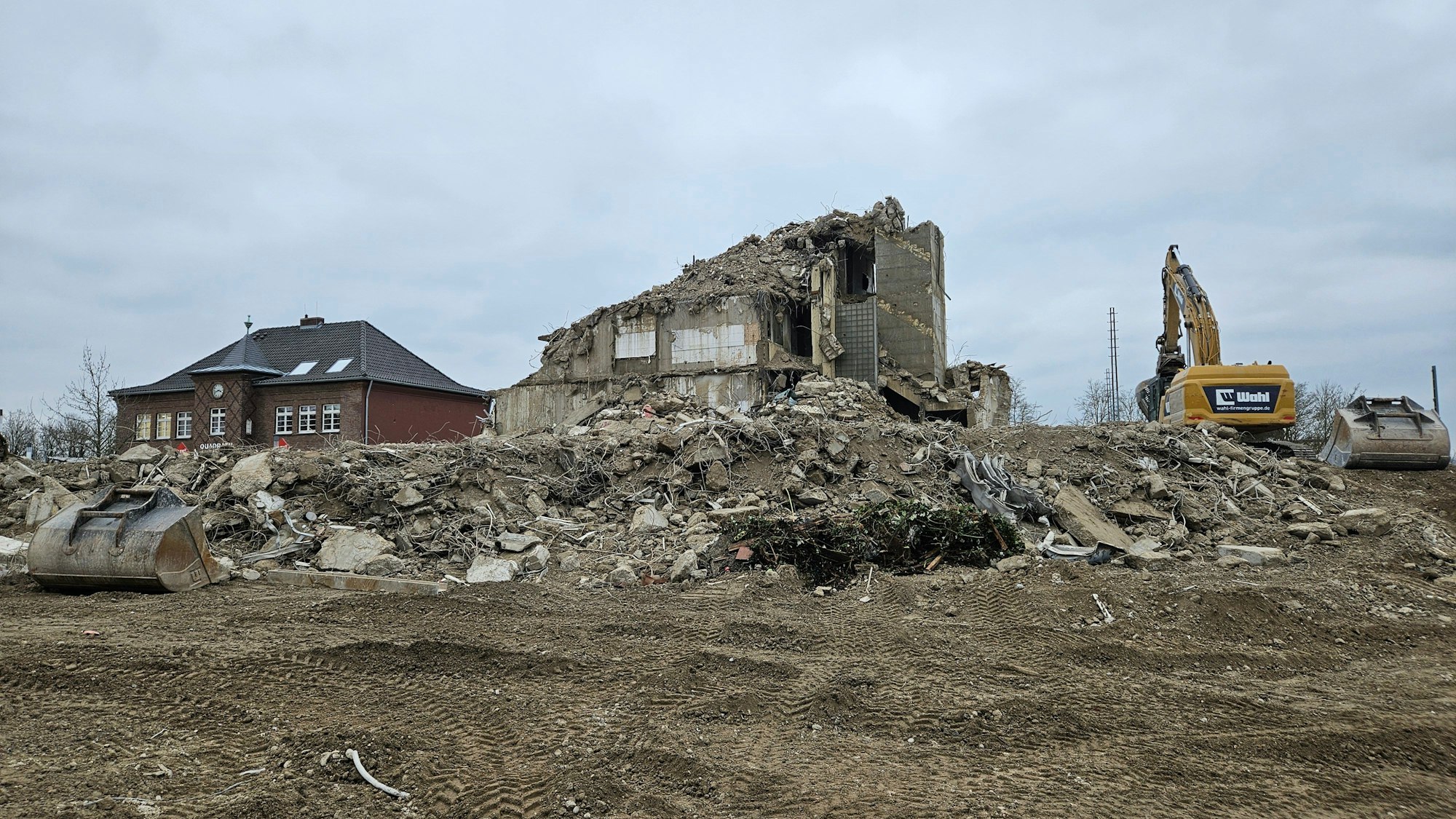 Zu sehen sind die Reste eines Hochhauses inmitten von Schutt, ein Bagger steht daneben.