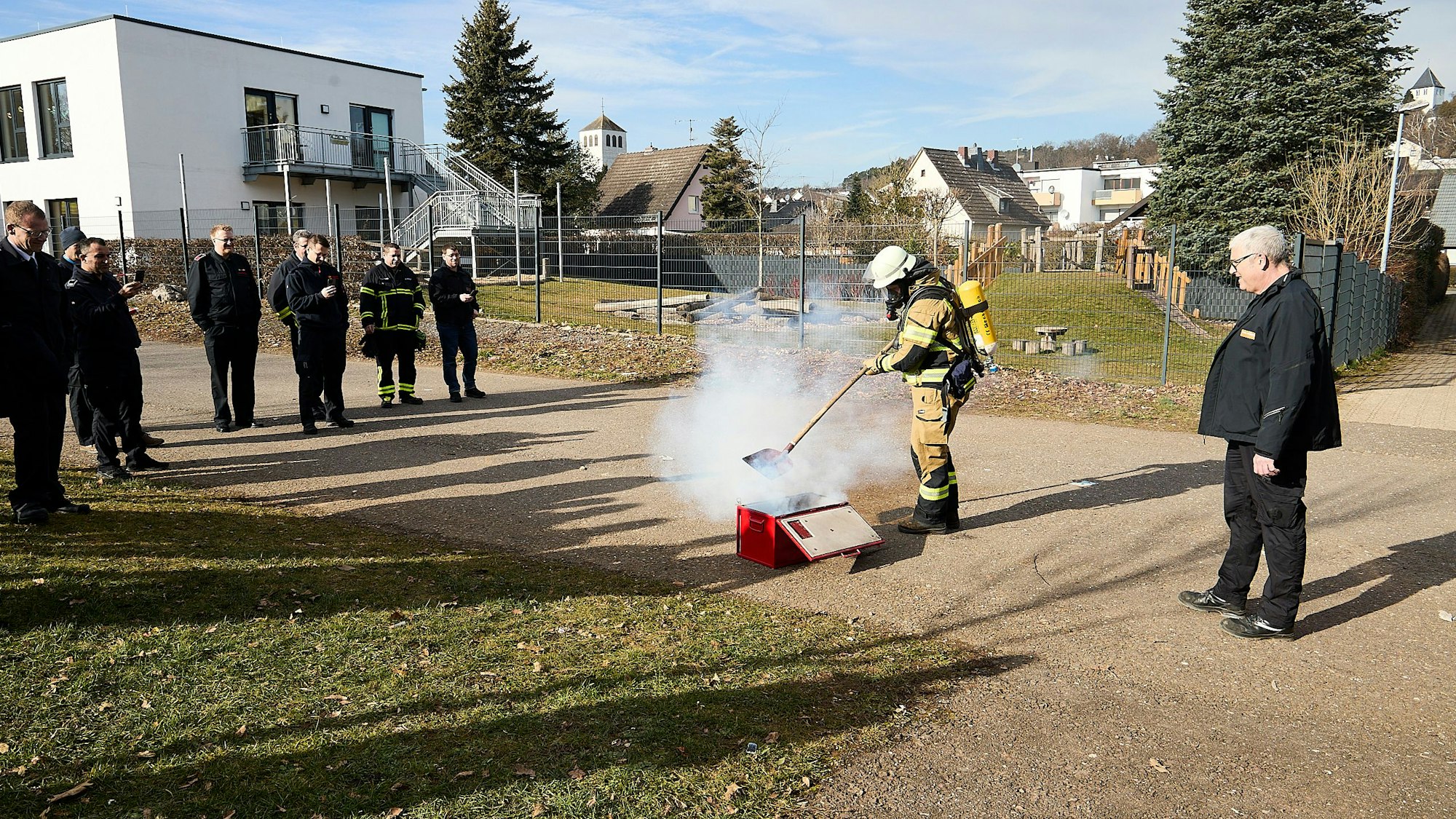 Feuerwehrleute stehen im Rahmen einer Vorführung um eine qualmende Kiste herum. Eine Einsatzkraft unter Atemschutz steht direkt daran und zeigt, wie die Feuerwehr vorgehen kann.