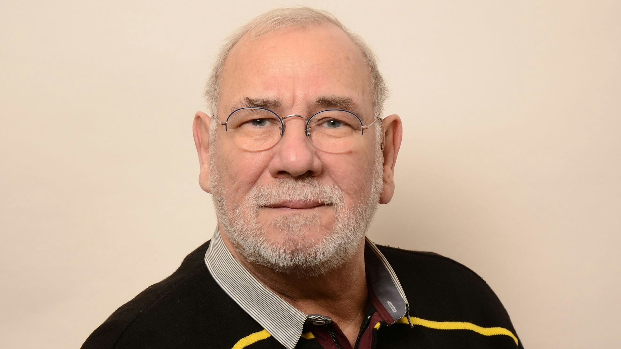 Ein älterer Mann trägt Brille und ein Hemd unter einem Pullover vor einem neutralen Hintergrund.