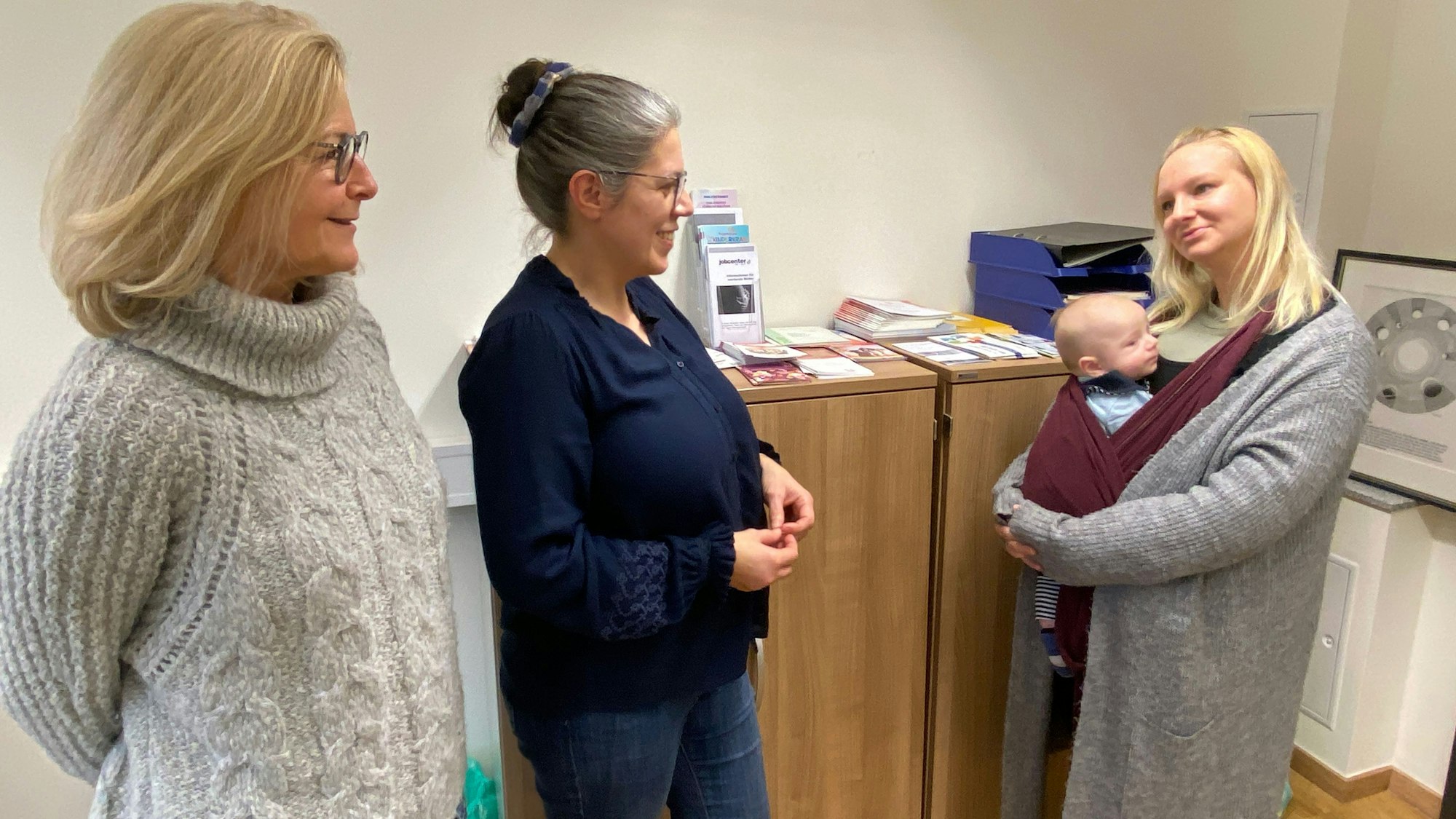 Drei Frauen stehen im Gespräch beieinander: Caritas-Fachbereichsleiterin Cilly von Sturm, Lotsin Birgit Hellmann und Laura Gardau mit Söhnchen Max im Tragetuch. Auf dem Büroschrank im Hintergrund liegen zahlreiche Broschüren und Flyer.
