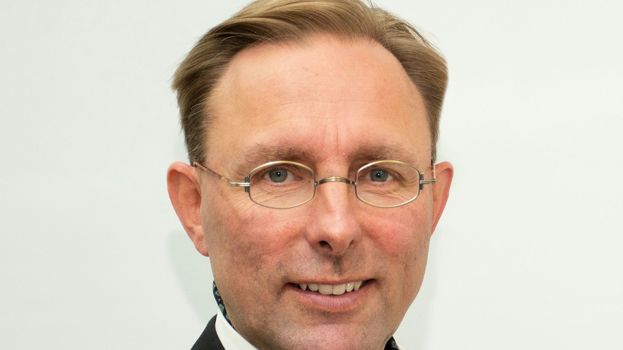 Ein Mann mit Brille im Anzug steht vor einem weißen Hintergrund und grinst.