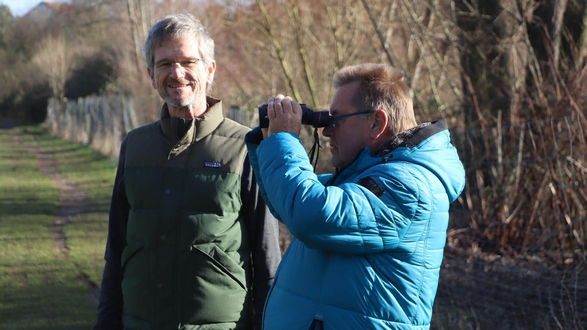 Zwei Männer stehen im Grünen, einer hält ein Fernglas.