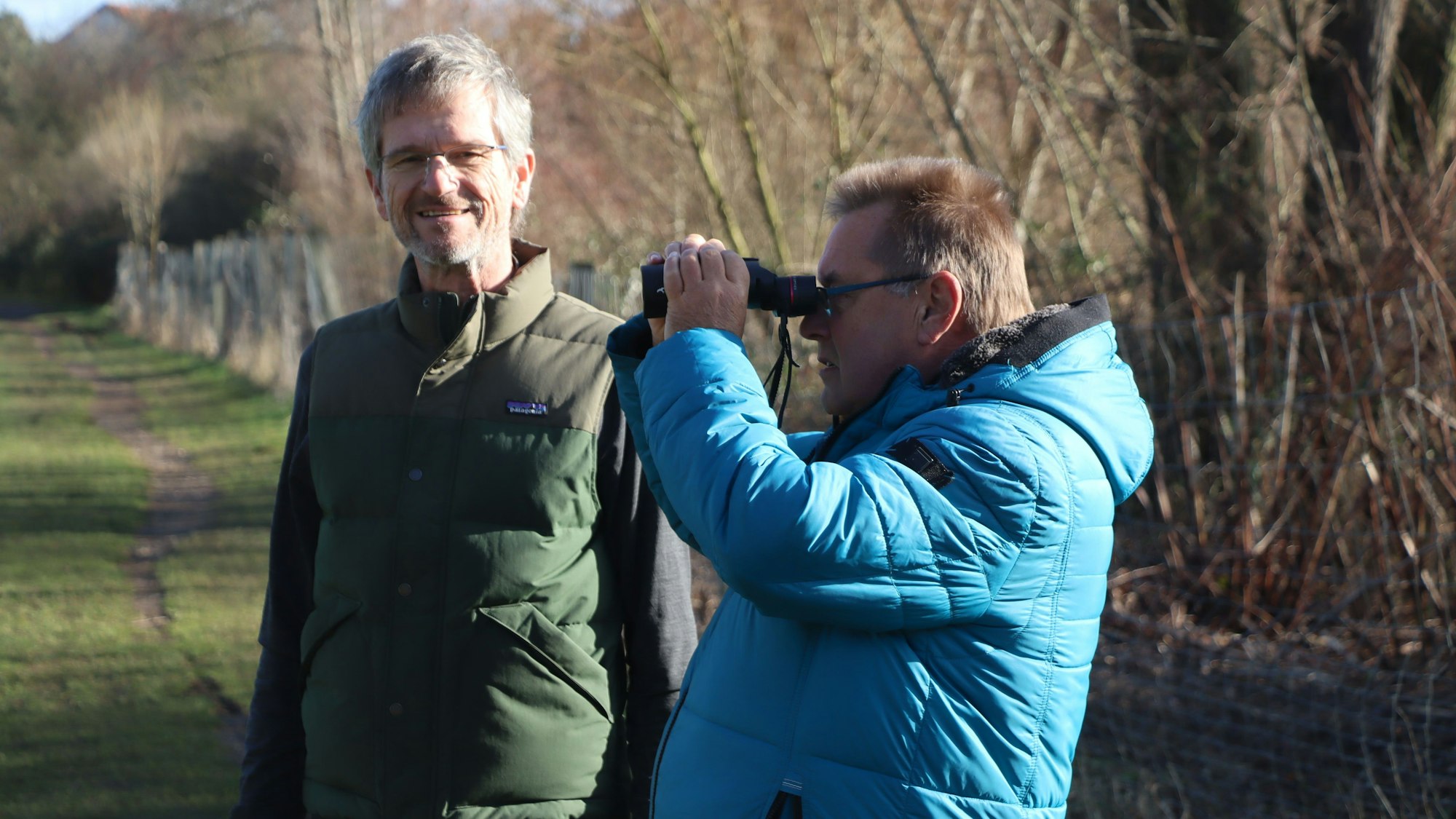 Zwei Männer stehen im Grünen, einer hält ein Fernglas.