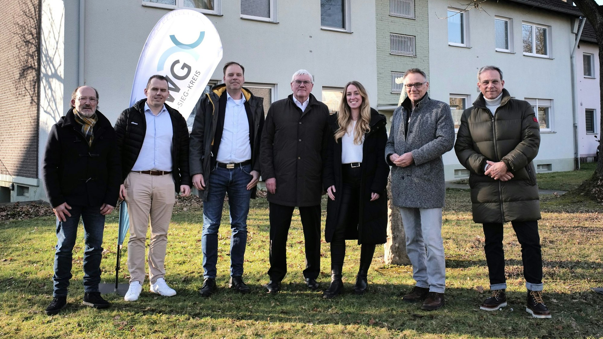 Sechs Männer und eine Frau stehen nebeneinander vor einem Mehrfamilienhaus.