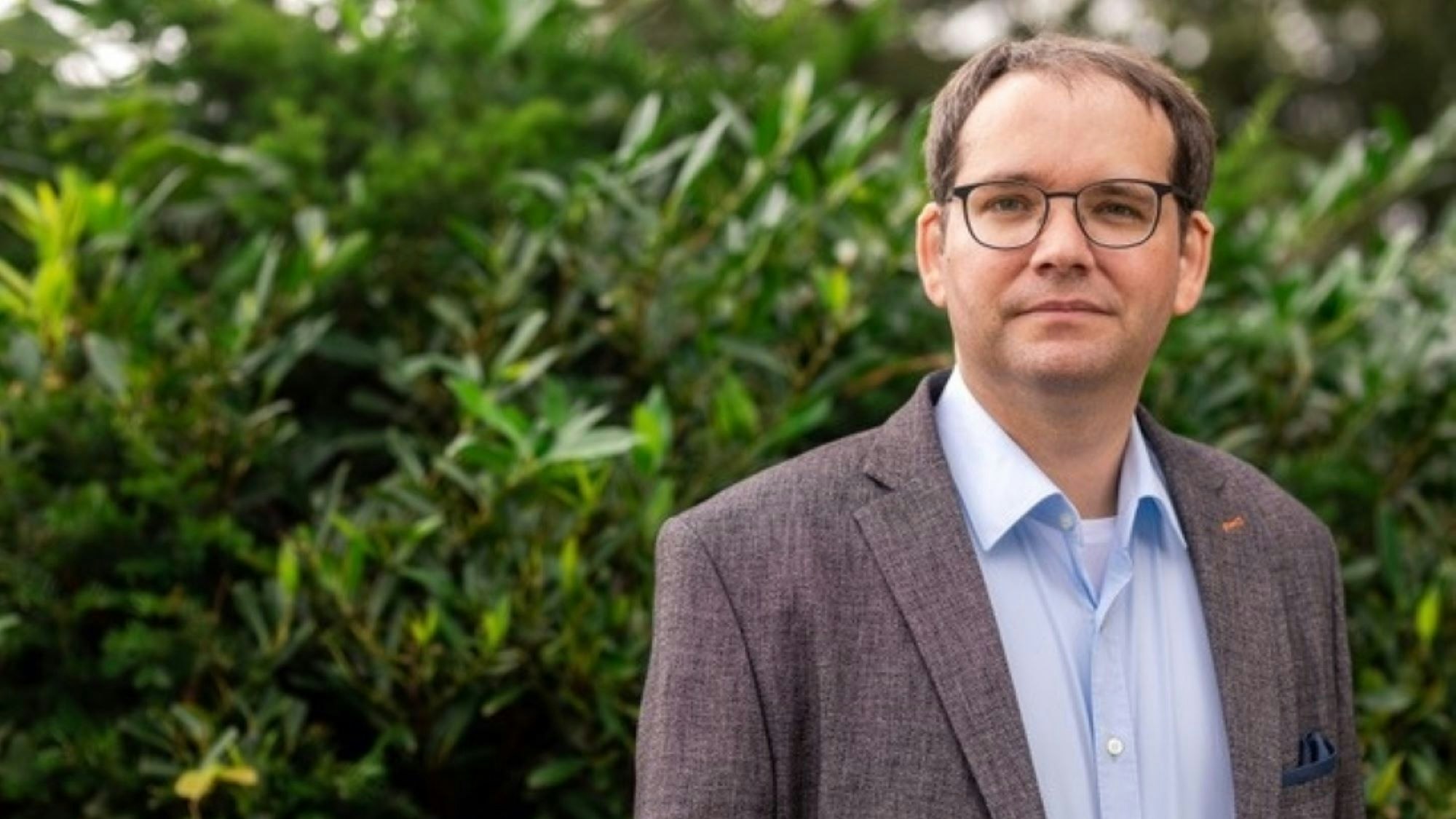 Ein Mann in Jackett, mit Brille steht vor einem grünen Busch.