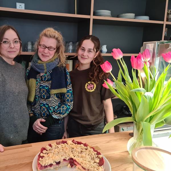 Drei Frauen stehen nebeneinander hinter einer Verkaufstheke, auf der ein Kirschstreusel-Kuchen und eine Vase mit Tulpen stehen.