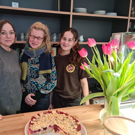 Drei Frauen stehen nebeneinander hinter einer Verkaufstheke, auf der ein Kirschstreusel-Kuchen und eine Vase mit Tulpen stehen.