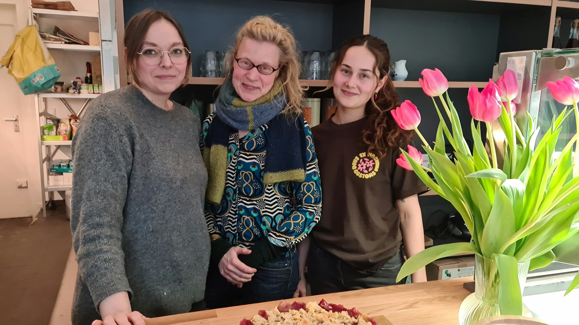 Drei Frauen stehen nebeneinander hinter einer Verkaufstheke, auf der ein Kirschstreusel-Kuchen und eine Vase mit Tulpen stehen.