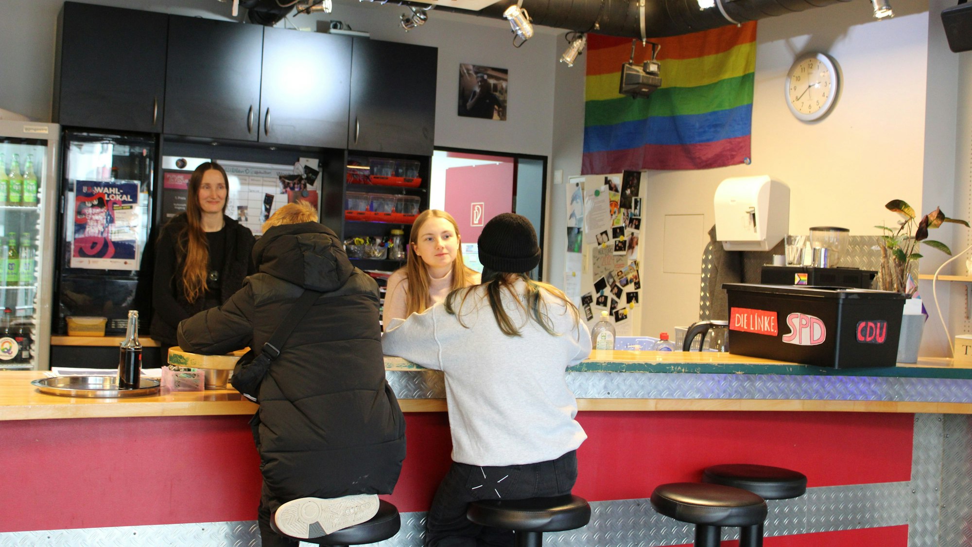 Zwei junge Leute sitzen an dem Tresen einer Bar, dahinter stehen zwei Frauen und unterhalten sich mit ihnen. Es hängt eine Regenbogenflagge an der Wand, die beschriebene selbstgebastelte Wahlurne steht auf dem Tresen.