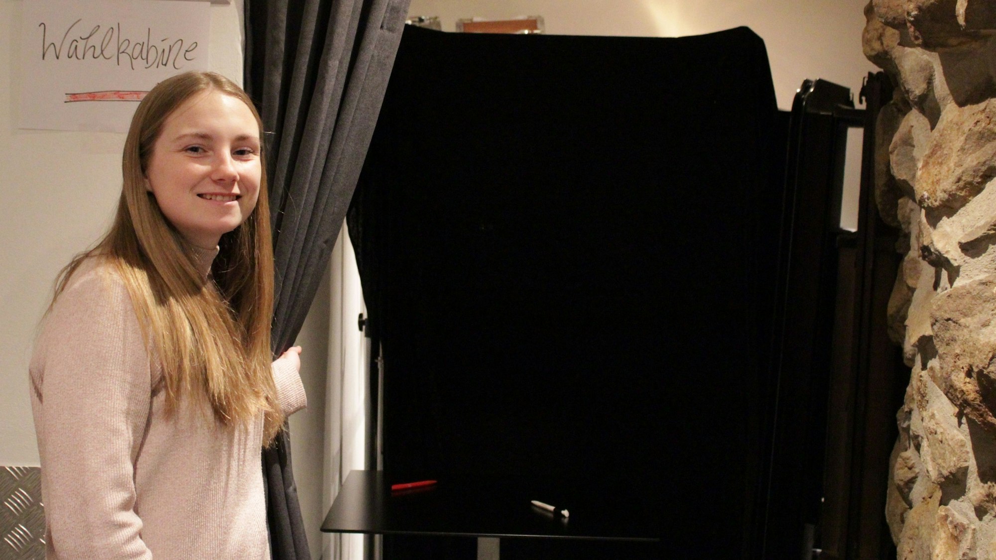 Eine junge Frau zieht einen Vorhang zur Seite, der den Blick auf einen Tisch freigibt, wo zwei Kugelschreiber liegen. Daneben steht ein Schild mit der Aufschrift „Wahlkabine“.