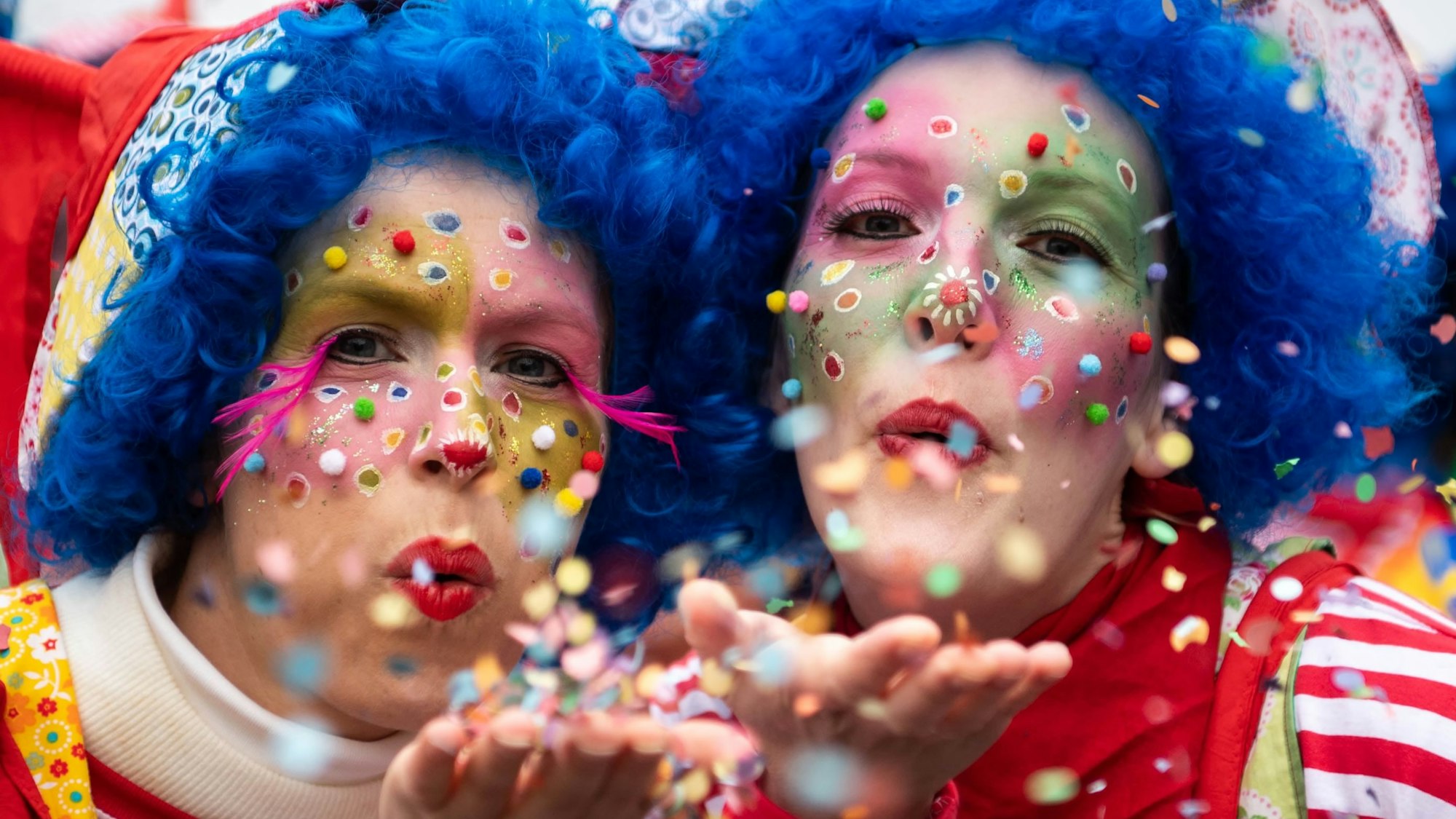 Das Bild zeigt zwei Frauen, die als Clowns kostümiert sind und von der Handfläche Konfetti in die Kamera pusten.