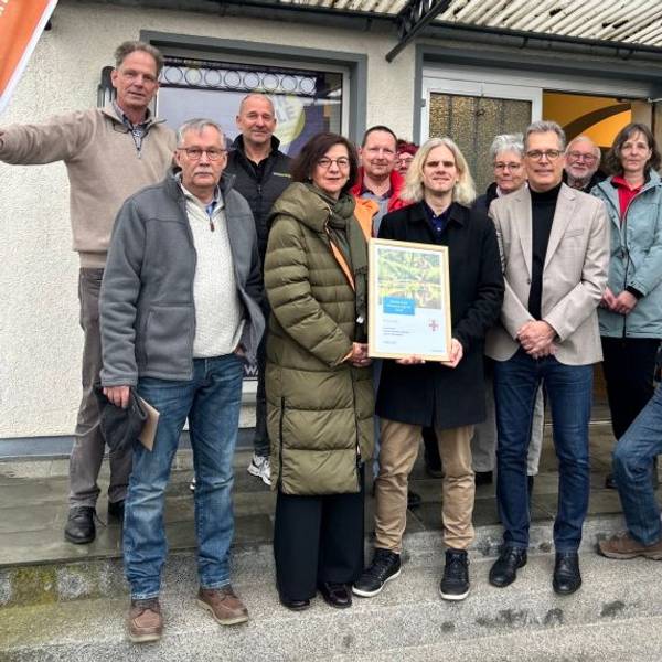 Eine Gruppe Menschen steht vor einem Haus und hält eine Urkunde in die Kamera.
