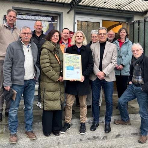 Eine Gruppe Menschen steht vor einem Haus und hält eine Urkunde in die Kamera.