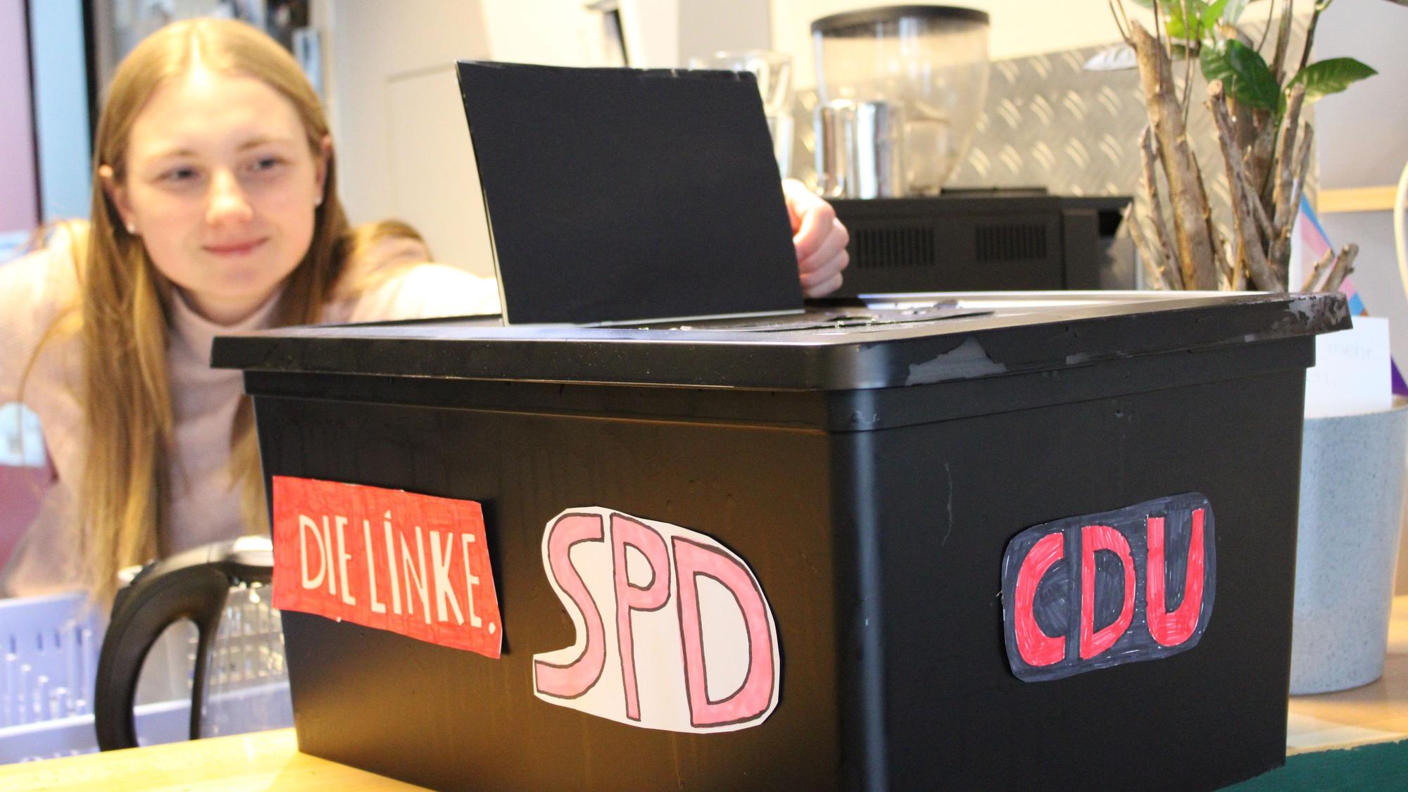Eine junge Frau öffnet eine Plastikkiste, die mit den Logos verschiedener Parteien beklebt ist: Die Logos der Linken, SPD und CDU sind sichtbar.