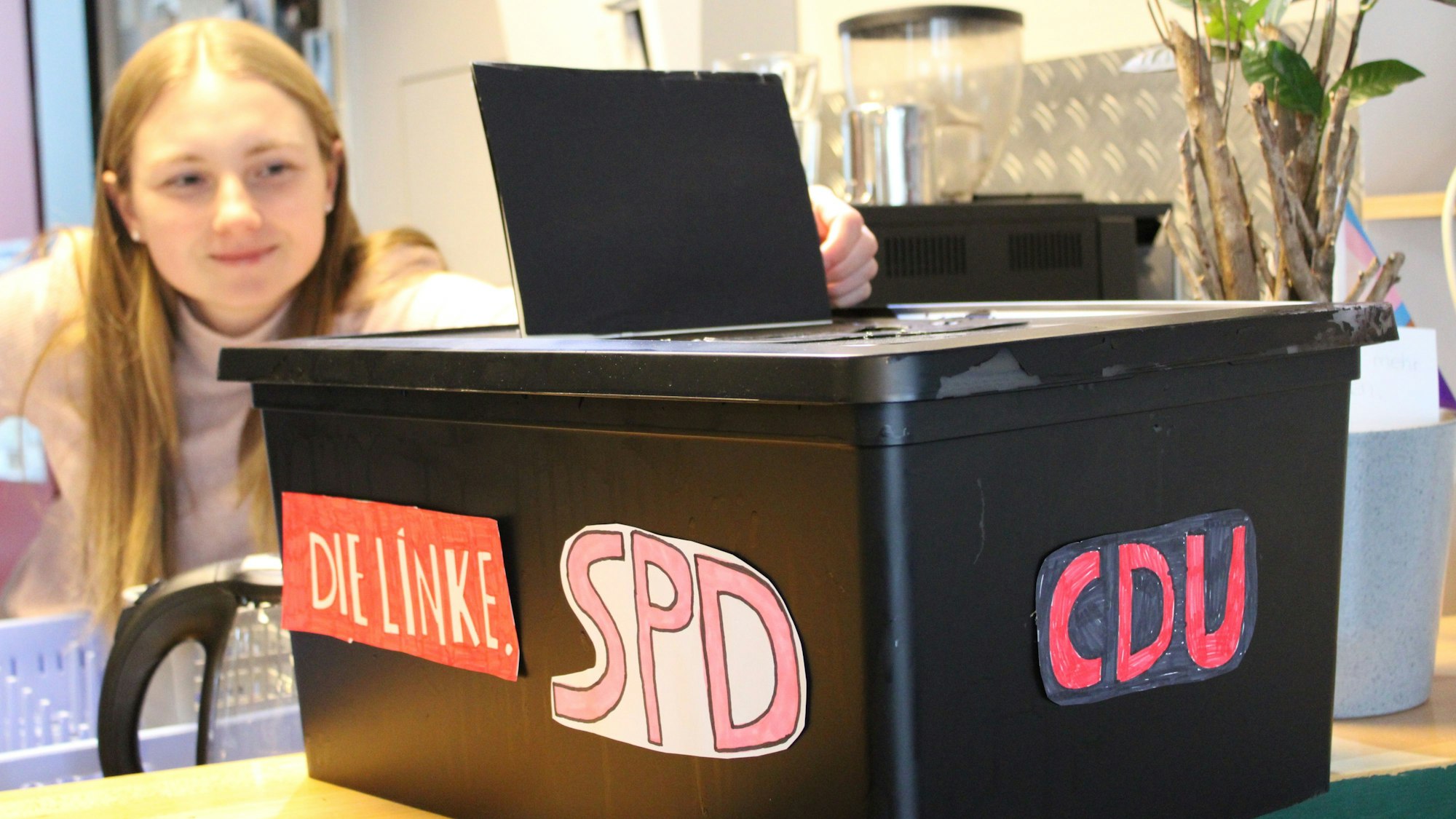 Eine junge Frau öffnet eine Plastikkiste, die mit den Logos verschiedener Parteien beklebt ist: Die Logos der Linken, SPD und CDU sind sichtbar.