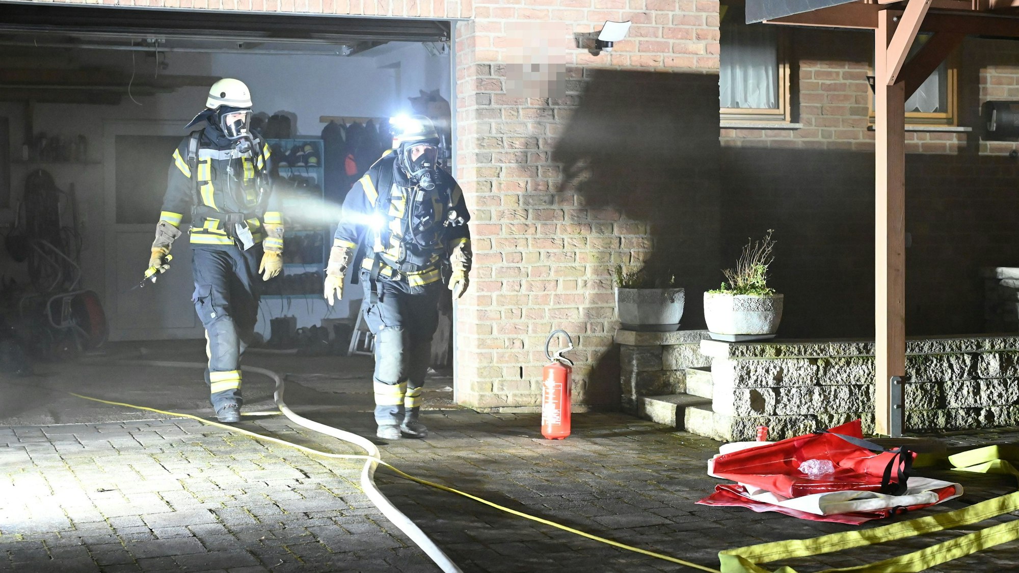 Zwei Feuerwehrleute mit Atemschutzausrüstung gehen aus der Garage eines Einfamilienhauses in Hellenthal-Eichen.