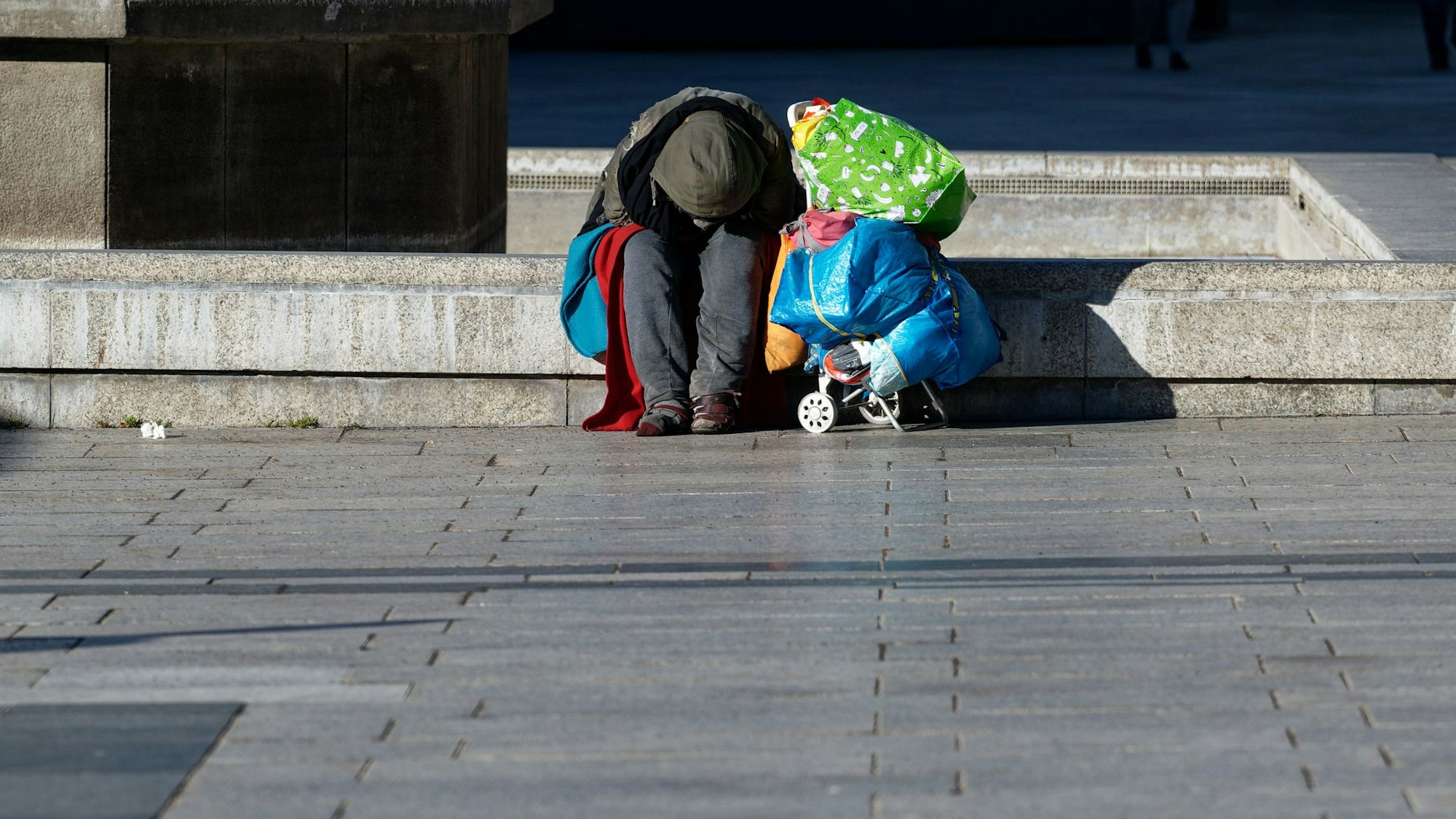 ARCHIV - 21.02.2021, Nordrhein-Westfalen, Köln: Ein Obdachloser wärmt sich auf dem Roncalliplatz vor dem Dom sitzend in der Sonne. (zu dpa: «Millionen armutsgefährdete NRW-Bürger - was könnte helfen?») Foto: Henning Kaiser/dpa +++ dpa-Bildfunk +++