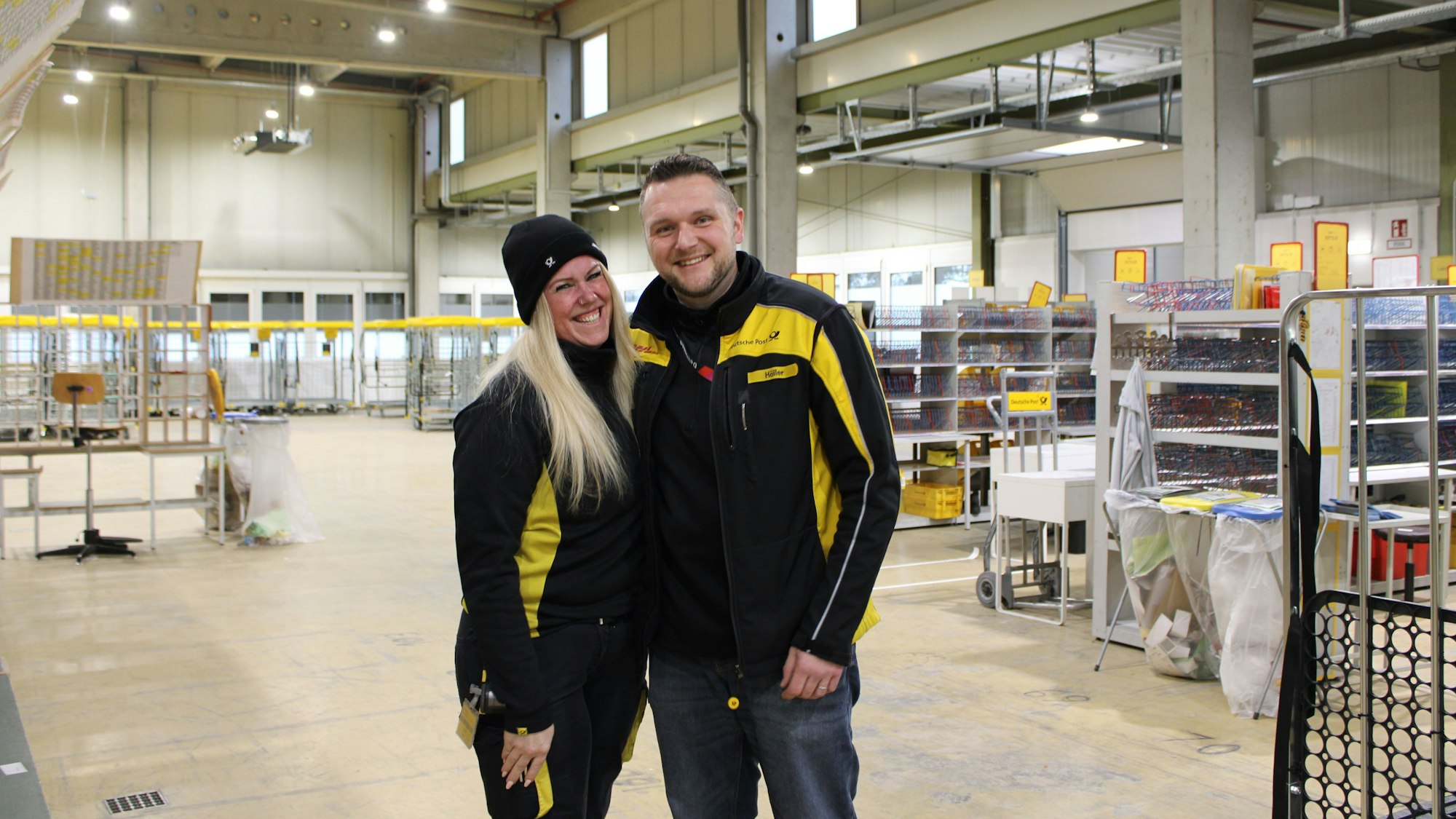 Eine Frau und ein Mann stehen Arm in Arm in der Halle des DHL-Zustellstützpunkts, beide tragen ihre Arbeitskleidung.