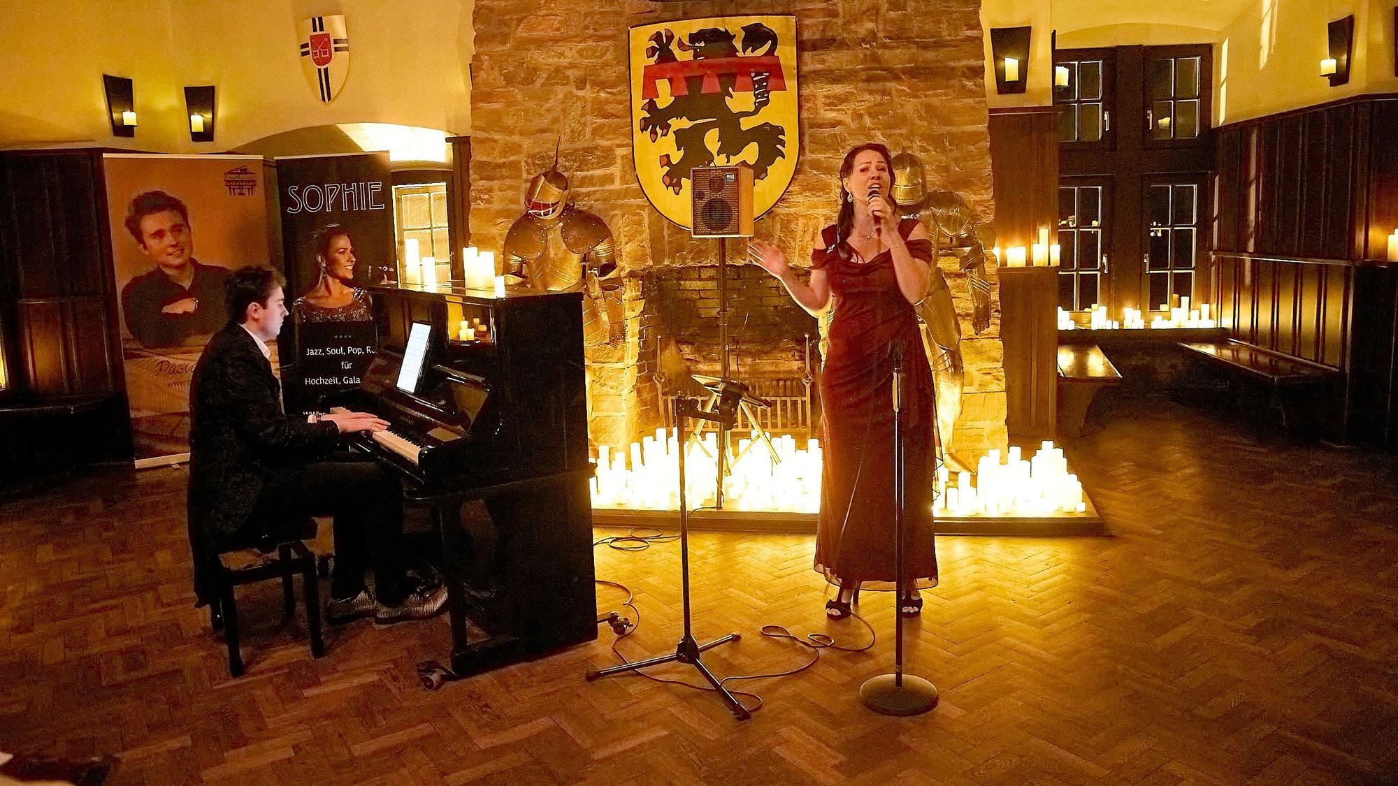 Eine Sängerin steht in einem langen roten Kleid vor einem mit vielen Kerzen beleuchteten Kamin. Neben dem Kamin stehen alte Ritterrüstungen und neben der Sängerin steht ein Klavier, an dem ein Mann sitzt und spielt.