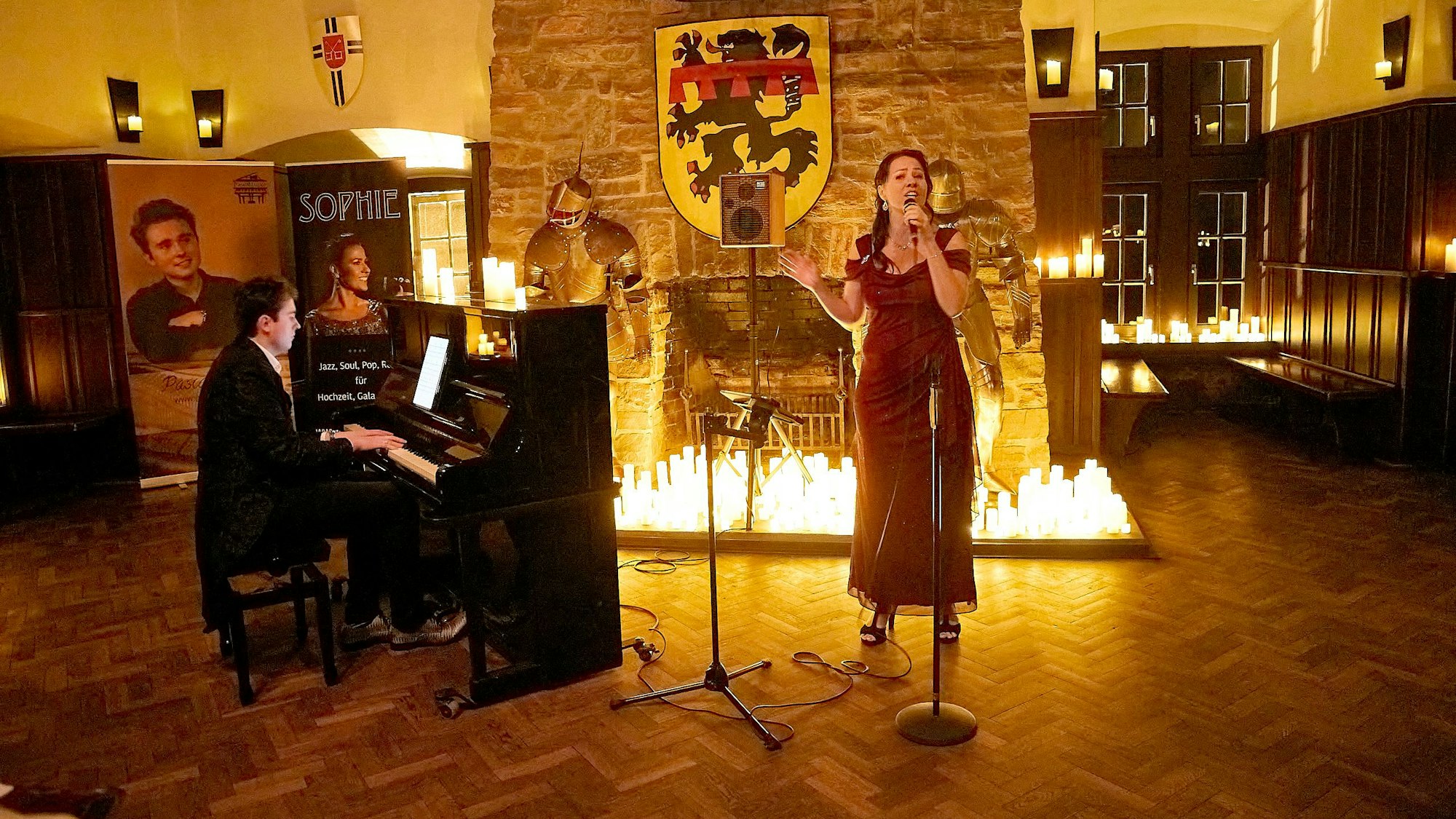 Eine Sängerin steht in einem langen roten Kleid vor einem mit vielen Kerzen beleuchteten Kamin. Neben dem Kamin stehen alte Ritterrüstungen und neben der Sängerin steht ein Klavier, an dem ein Mann sitzt und spielt.