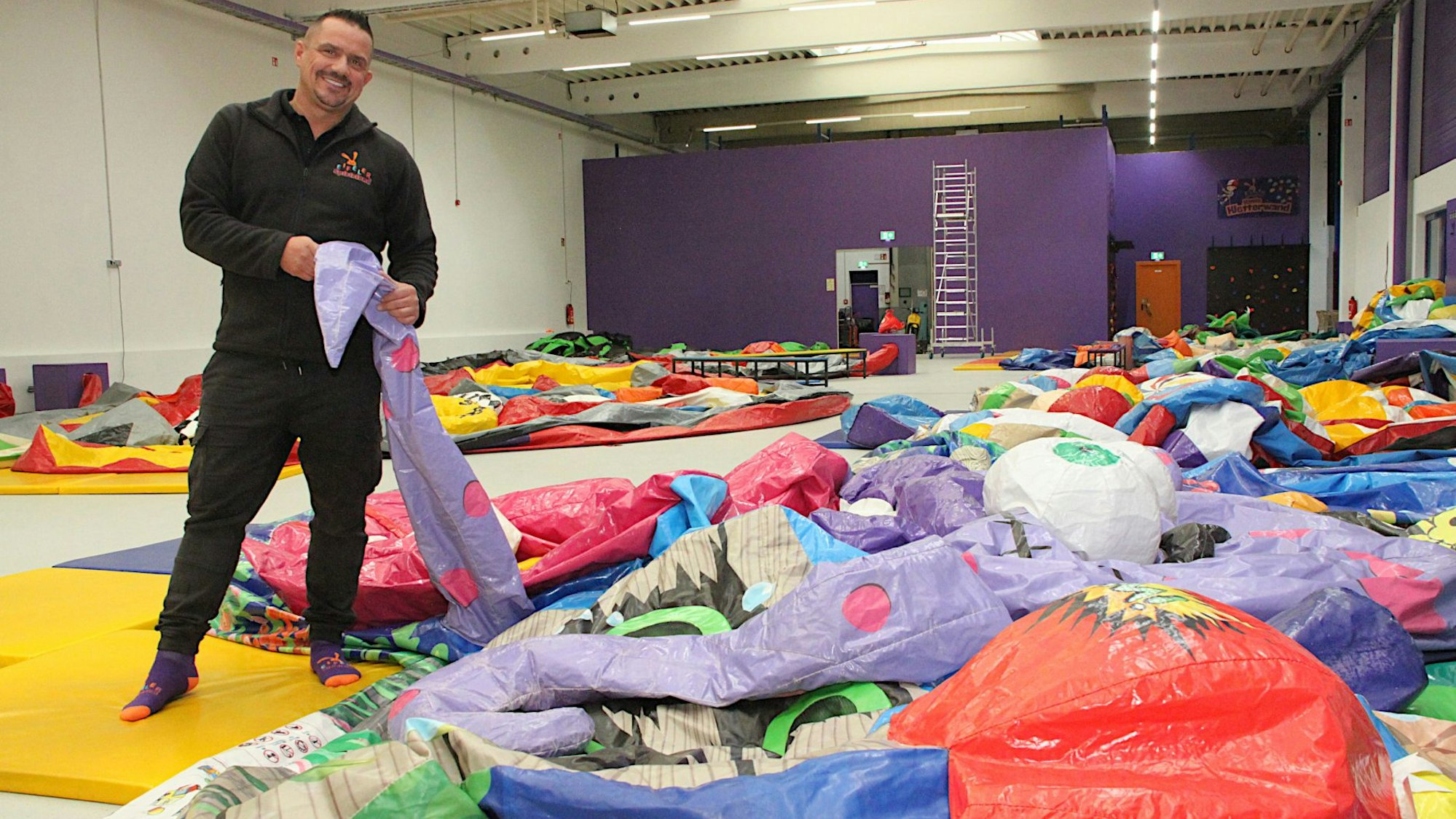Ein Mann im schwarzen Pullover steht in einer Halle und hält eine bunte Kunststoffbahn in den Händen. Sie gehört zu einer Hüpfburg für Kinder, die noch nicht aufgeblasen ist.