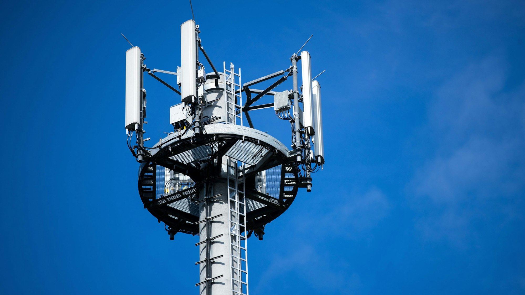 Ein Mast mit verschiedenen Antennen von Mobilfunkanbietern steht vor blauem Himmel.