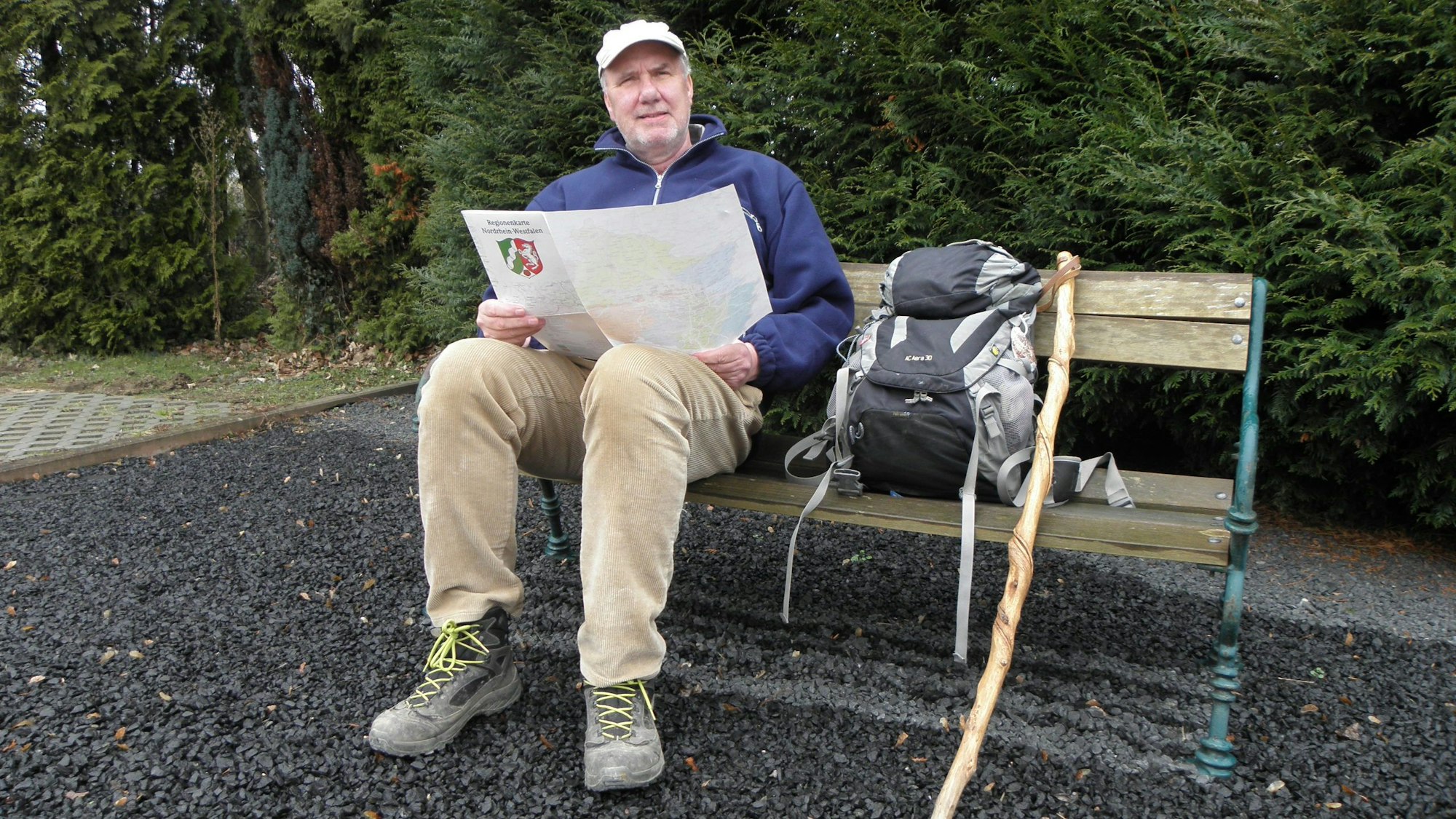 Ralf Spilles sitzt auf einer Bank. Er hält eine Karte mit dem NRW-Wappen in der Hand. Neben ihm stehen sein Wanderrucksack und ein Stock.