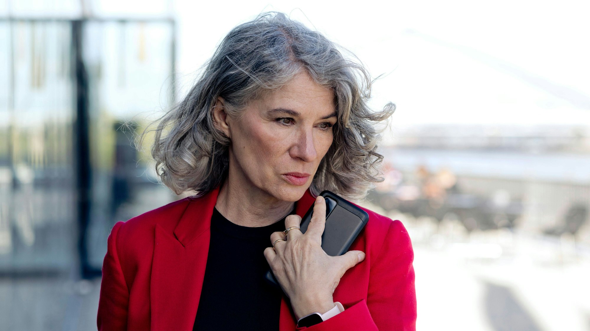 Frau mit grauen Haaren hält ihr Smartphone an ihre Schulter und blickt schräg nach unten ins Leere