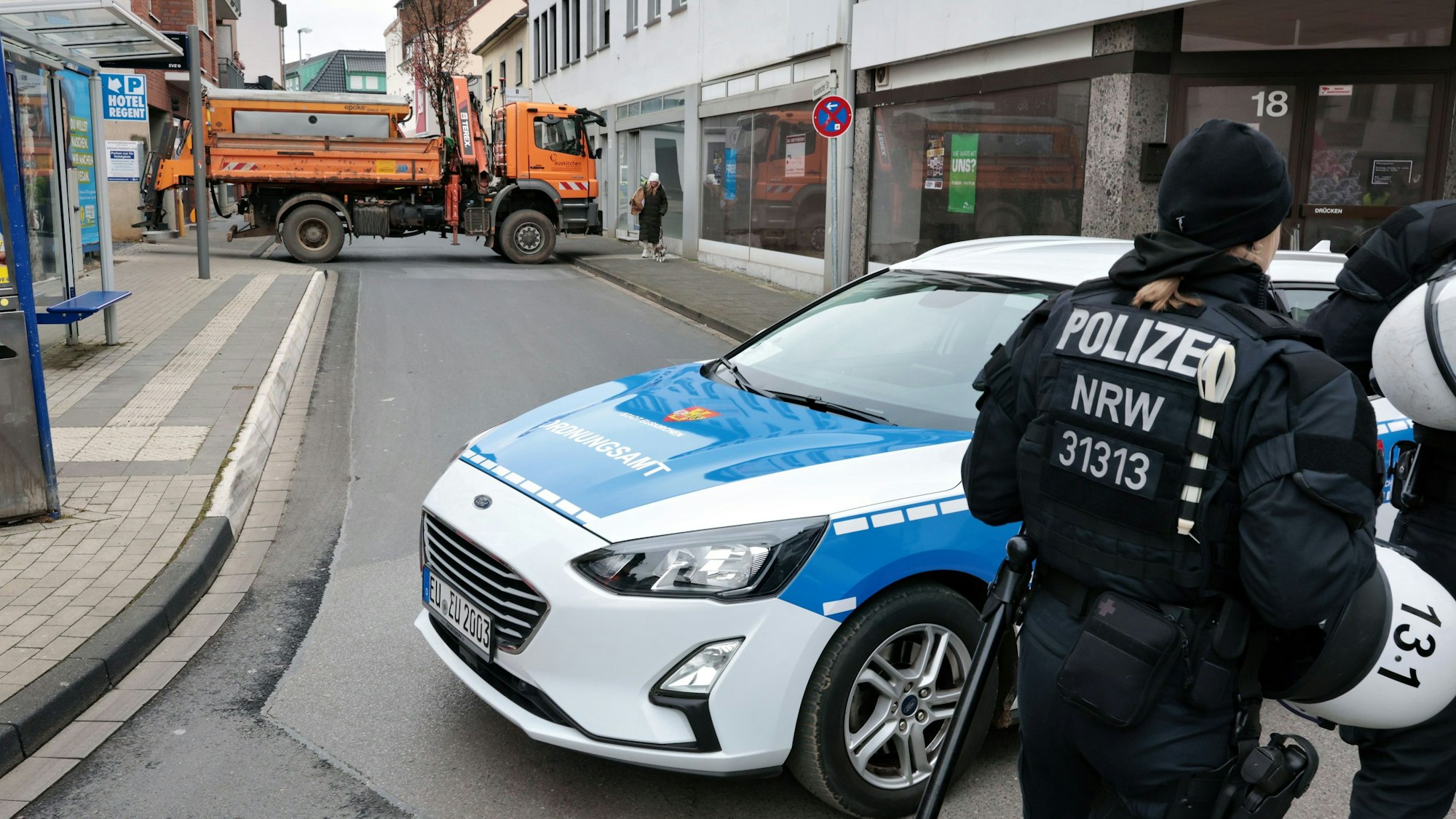 Eine Straße in Euskirchen wird durch ein blau-weißes Fahrzeug und einen orangen Lastwagen der Stadt blockiert. Von hinten ist eine Polizistin zu sehen, die mit ihren Kollegen die politischen Veranstaltungen sichert.