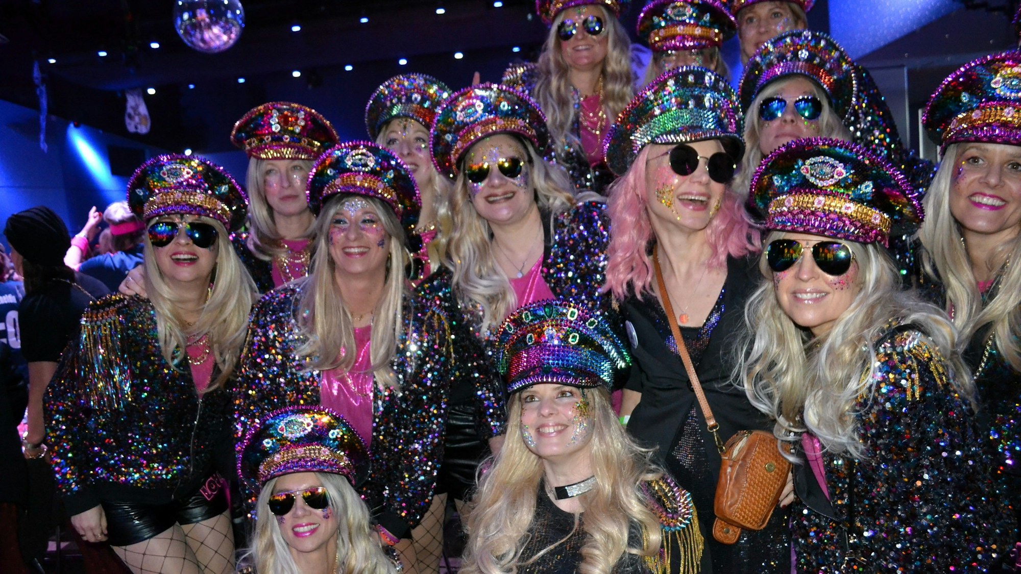 Eine Gruppe von Frauen mit blonden Perücken, glitzernden Jacken und Mützen. Einige tragebn Sonnenbrillen.