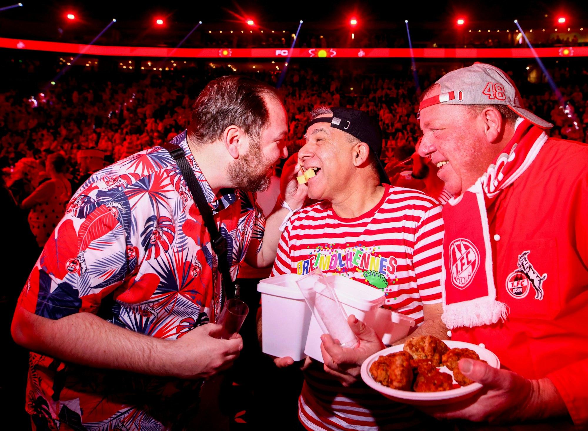 Mann füttert anderen Mann mit Käsewürfeln, weiterer Mann daneben hält einen Teller mit Frikadellen in der Hand.