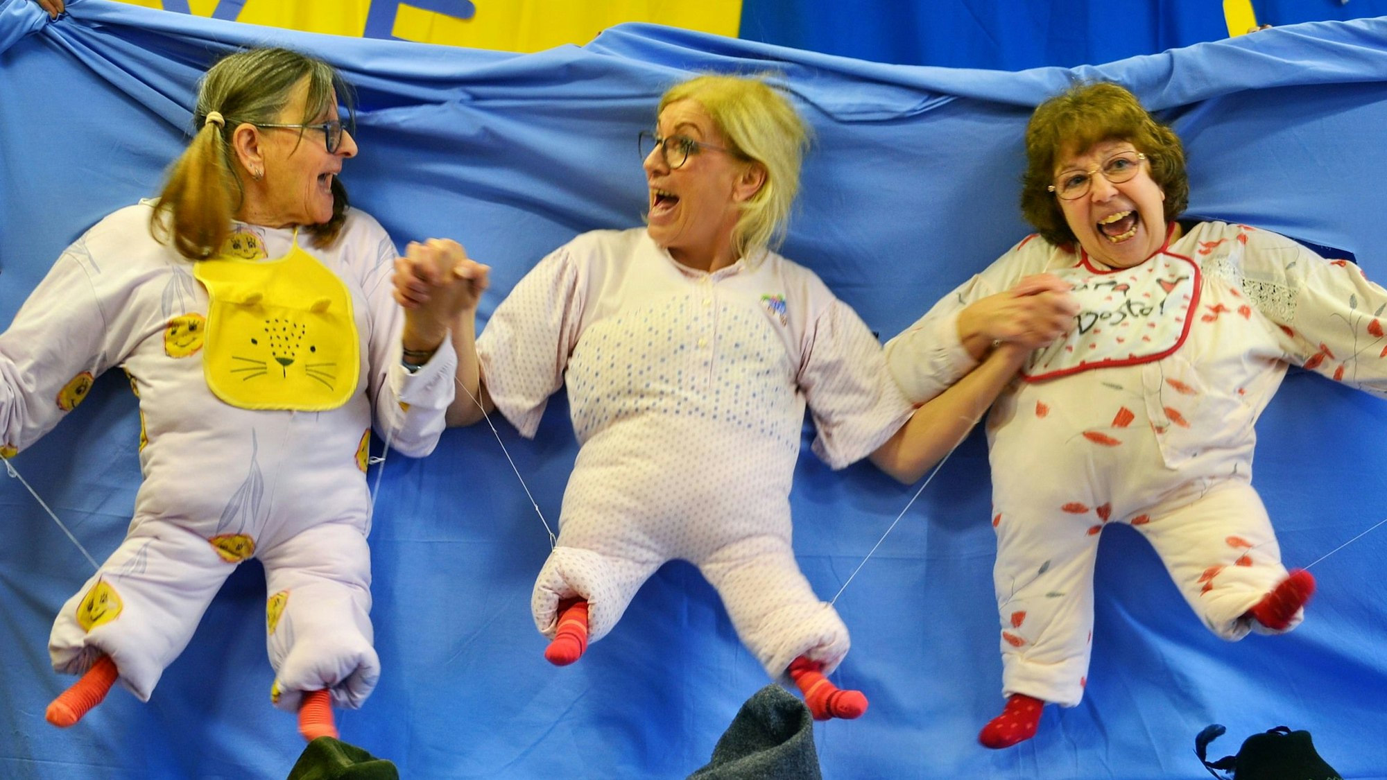 Drei der Jecken Wiewer haben sich in Babys verwandelt: Ihre Köpfe und Arme haben sie durch ein blaues Tuch gesteckt, auf dem kleine ausgestopfte Strampler angebracht sind.