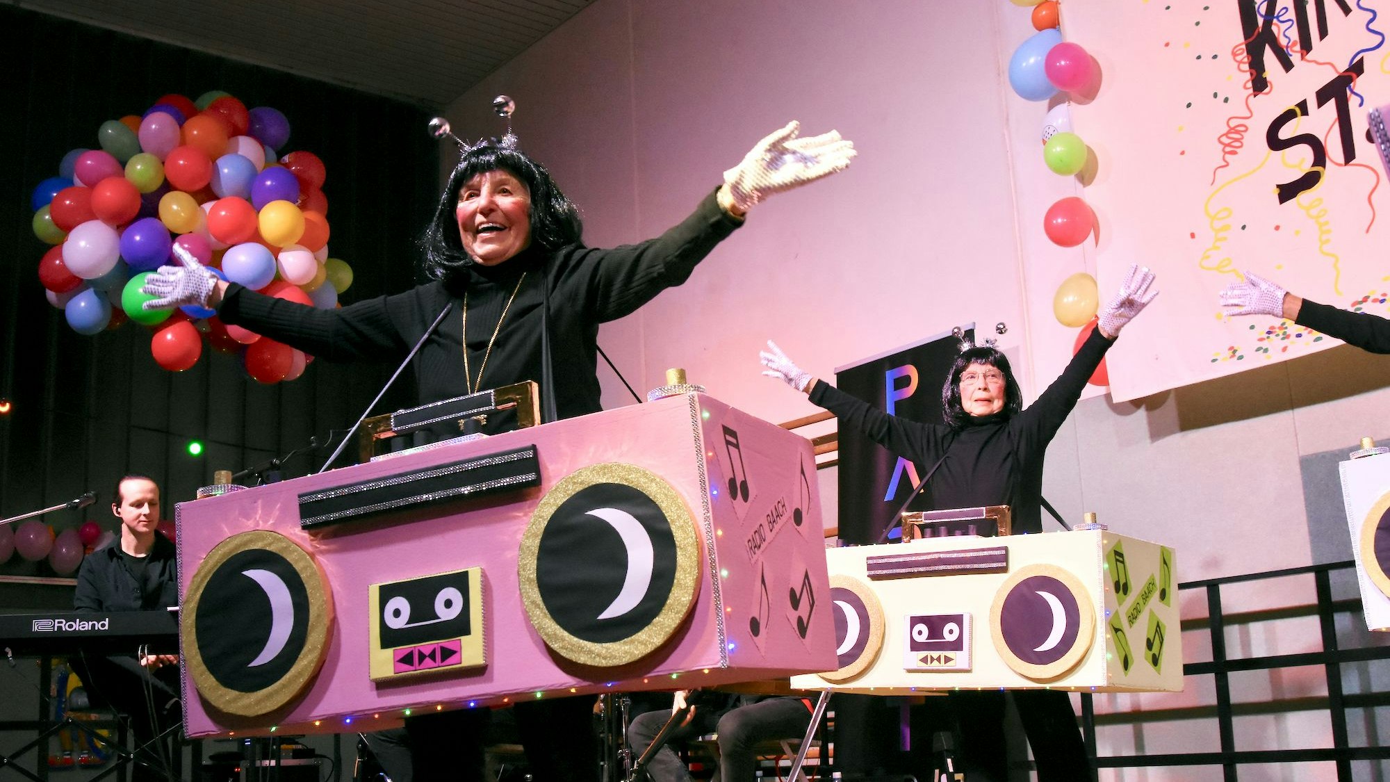 Zwei Frauen, mit großen Pappkartons als Radios kostümiert, tanzen auf einer Karnevalsbühne.