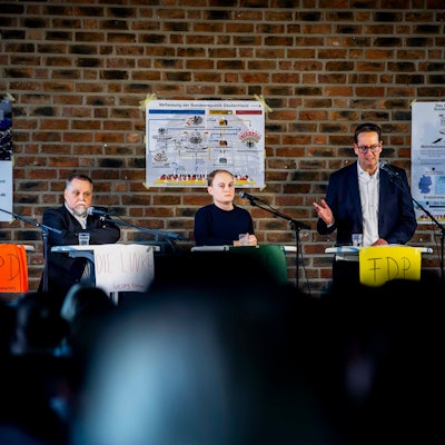 Eine Frau und vier Männer stehen jeweils hinter einem Tisch, vor sich Mikrofone.