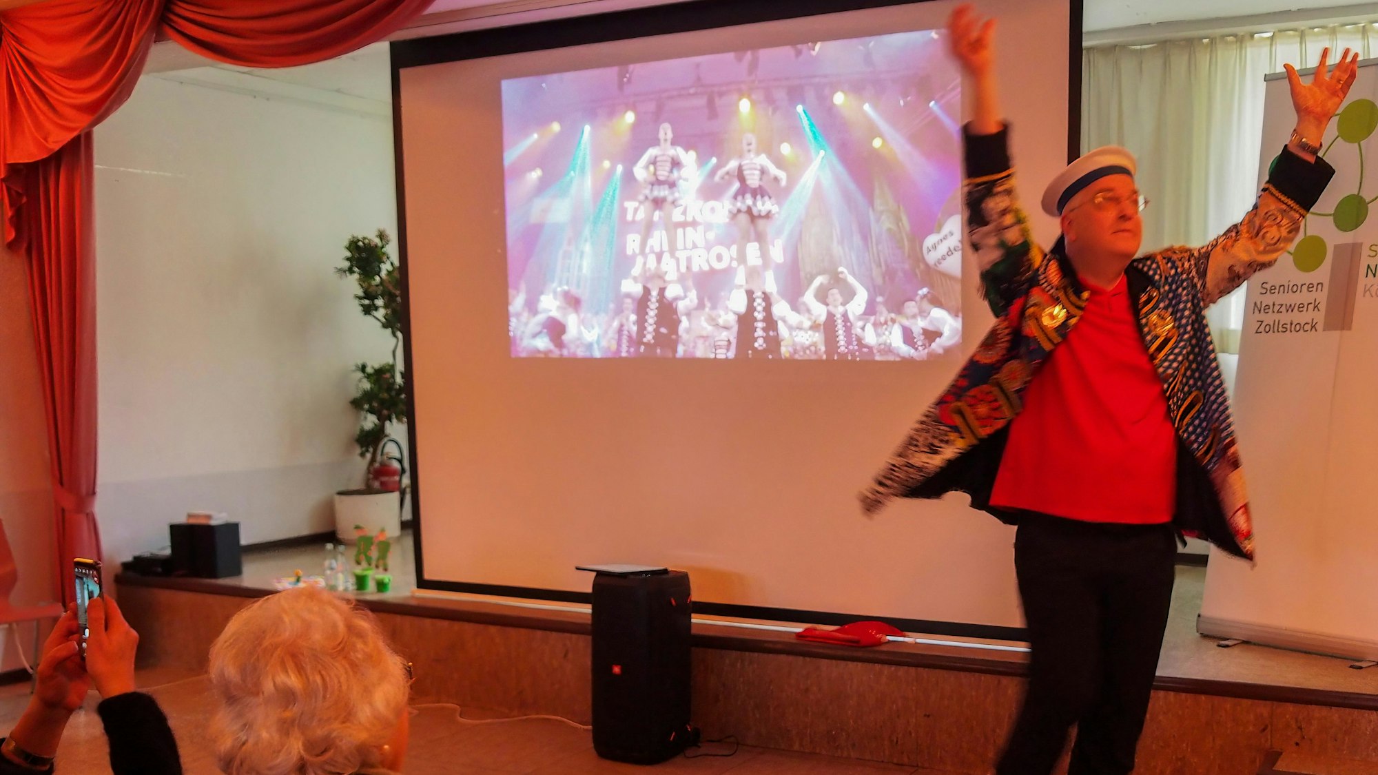 Uli Kievernagel steht mit erhobenen Armen auf der Bühne, hinter ihm auf einer Leinwand ist ein Tanzcorps zu sehen.